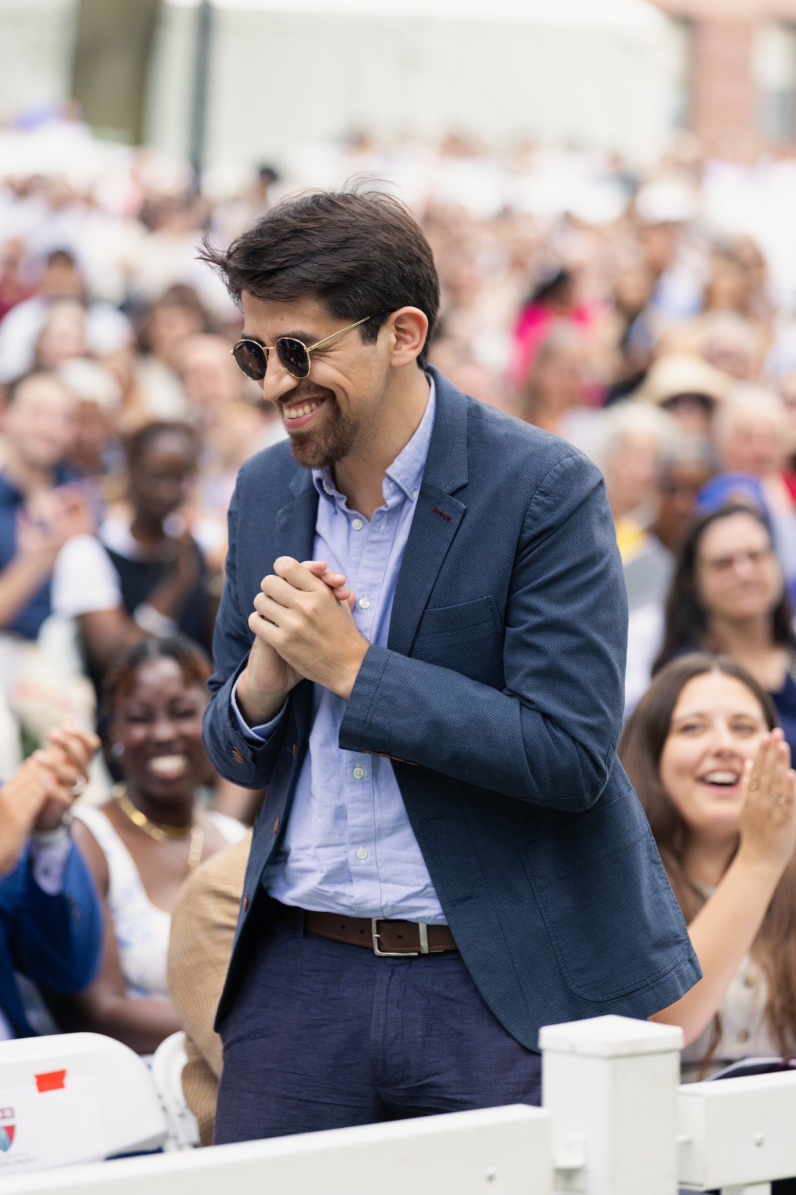 A person in a blue blazer and sunglasses smiles while clapping at an outdoor event, surrounded by a cheering crowd.
