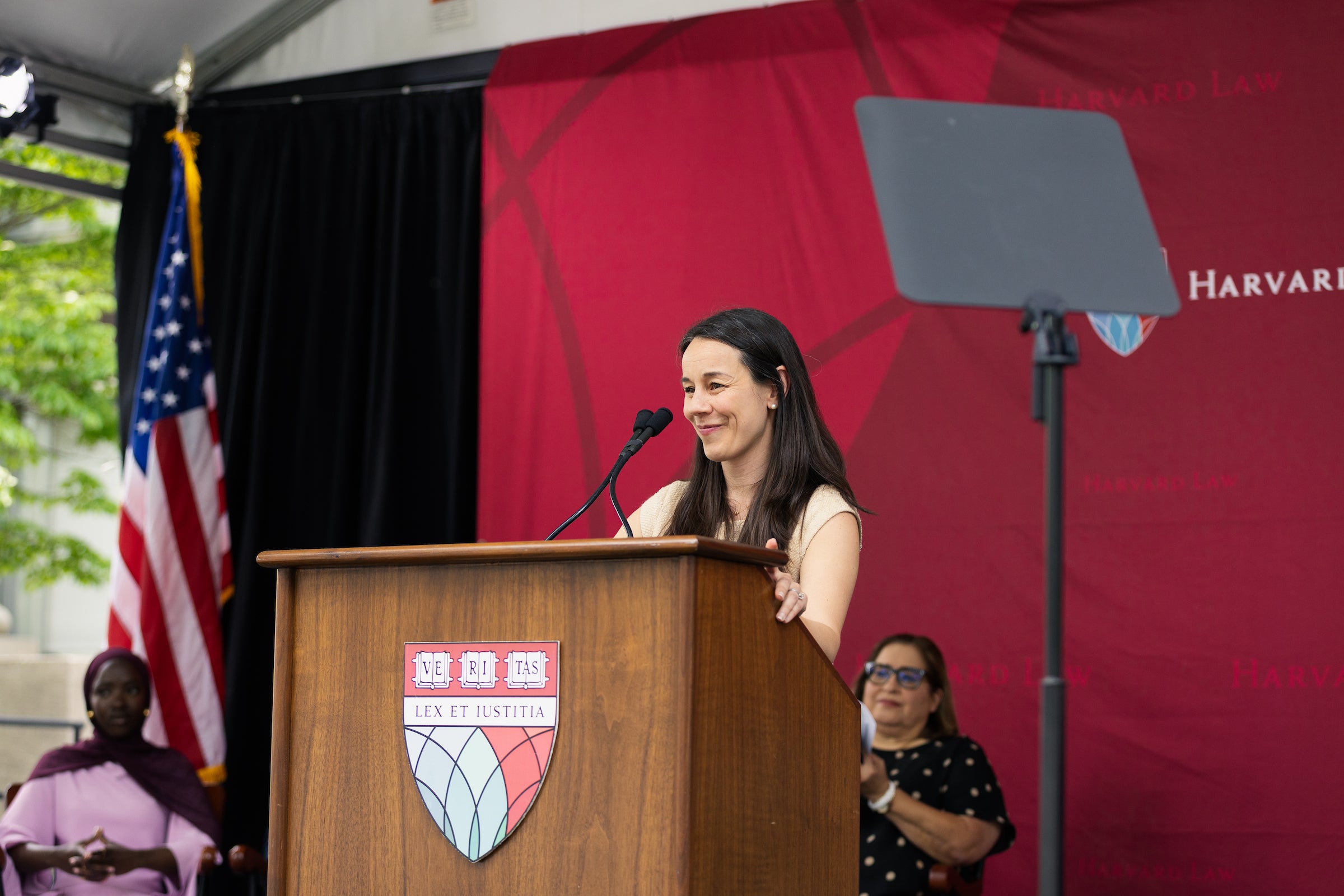 A person stands at a wooden podium with a Harvard Law shield, speaking into a microphone under an outdoor tent, with an American flag and a red backdrop behind them.