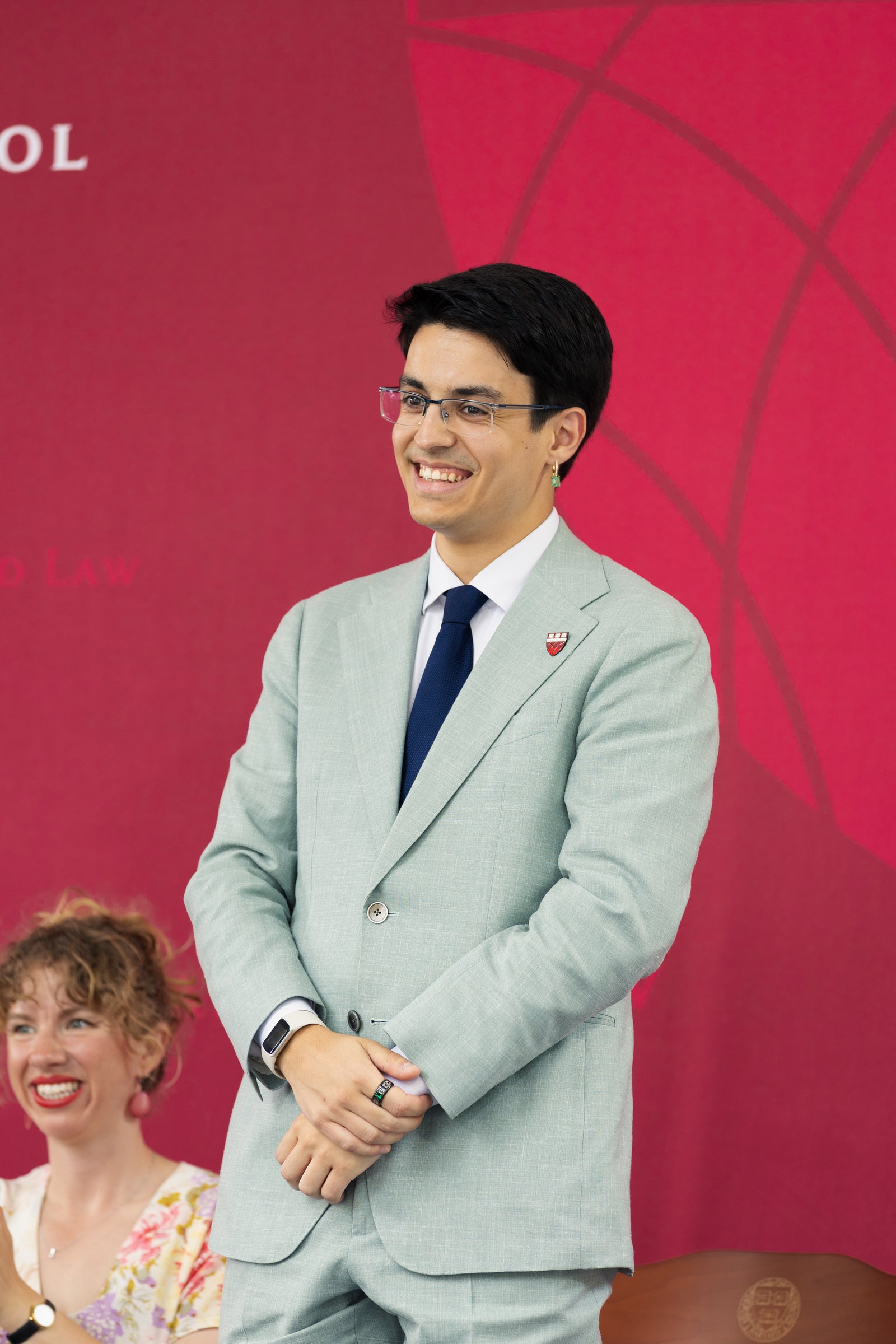 A person in a light gray suit and glasses smiles while standing in front of a red backdrop.