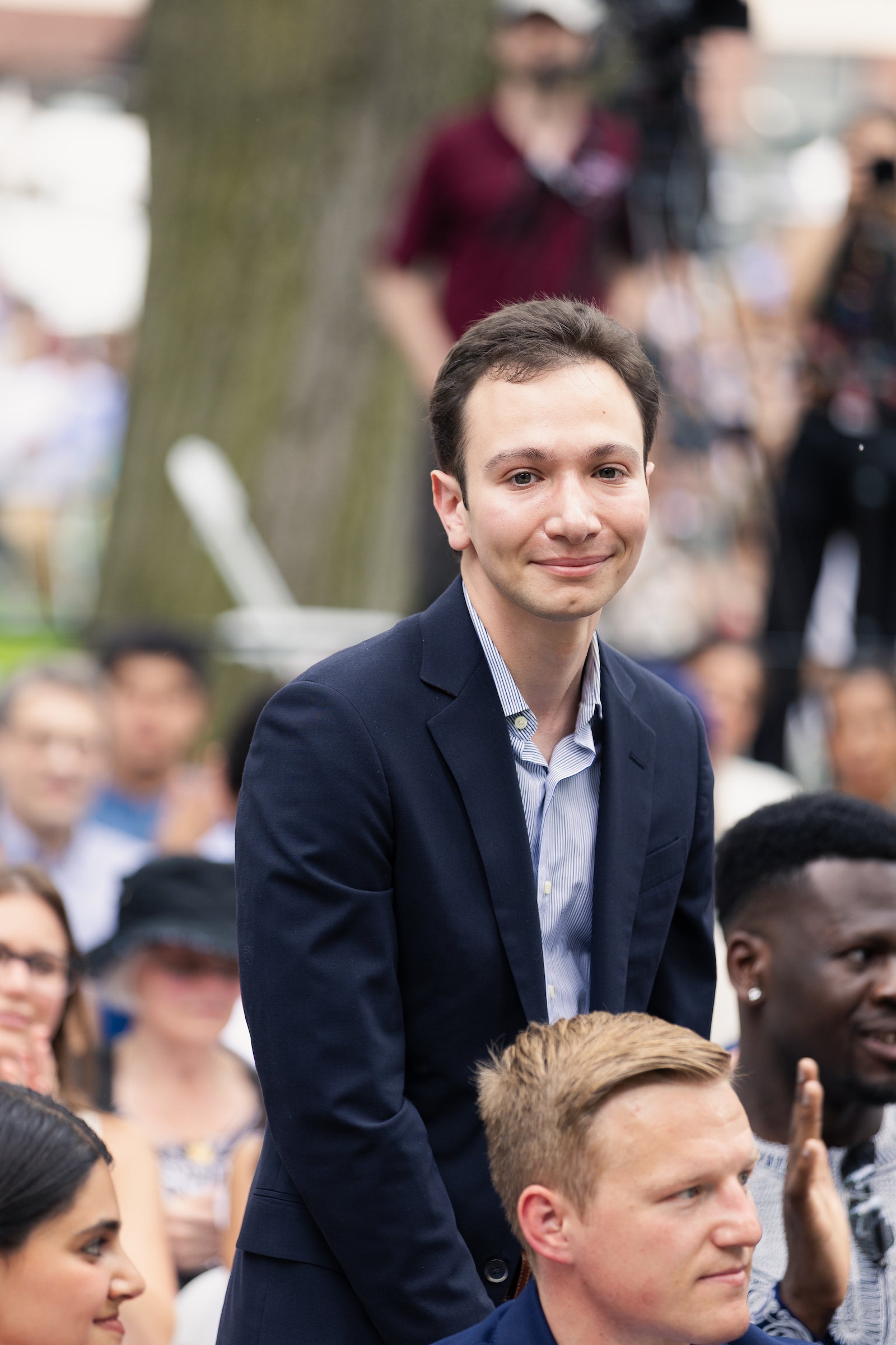 A person in a dark suit and light blue shirt smiles while standing among a crowd at an outdoor event.