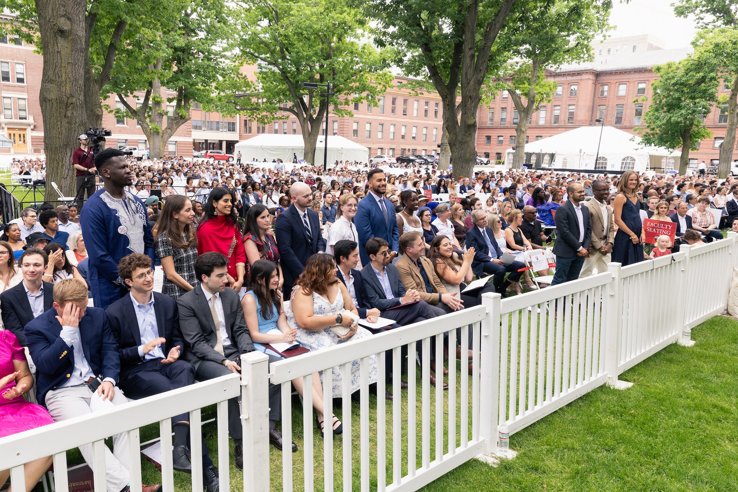 A large crowd gathers outdoors under trees for an event, with a row of graduates standing up as the crowd gives them a round of applause.