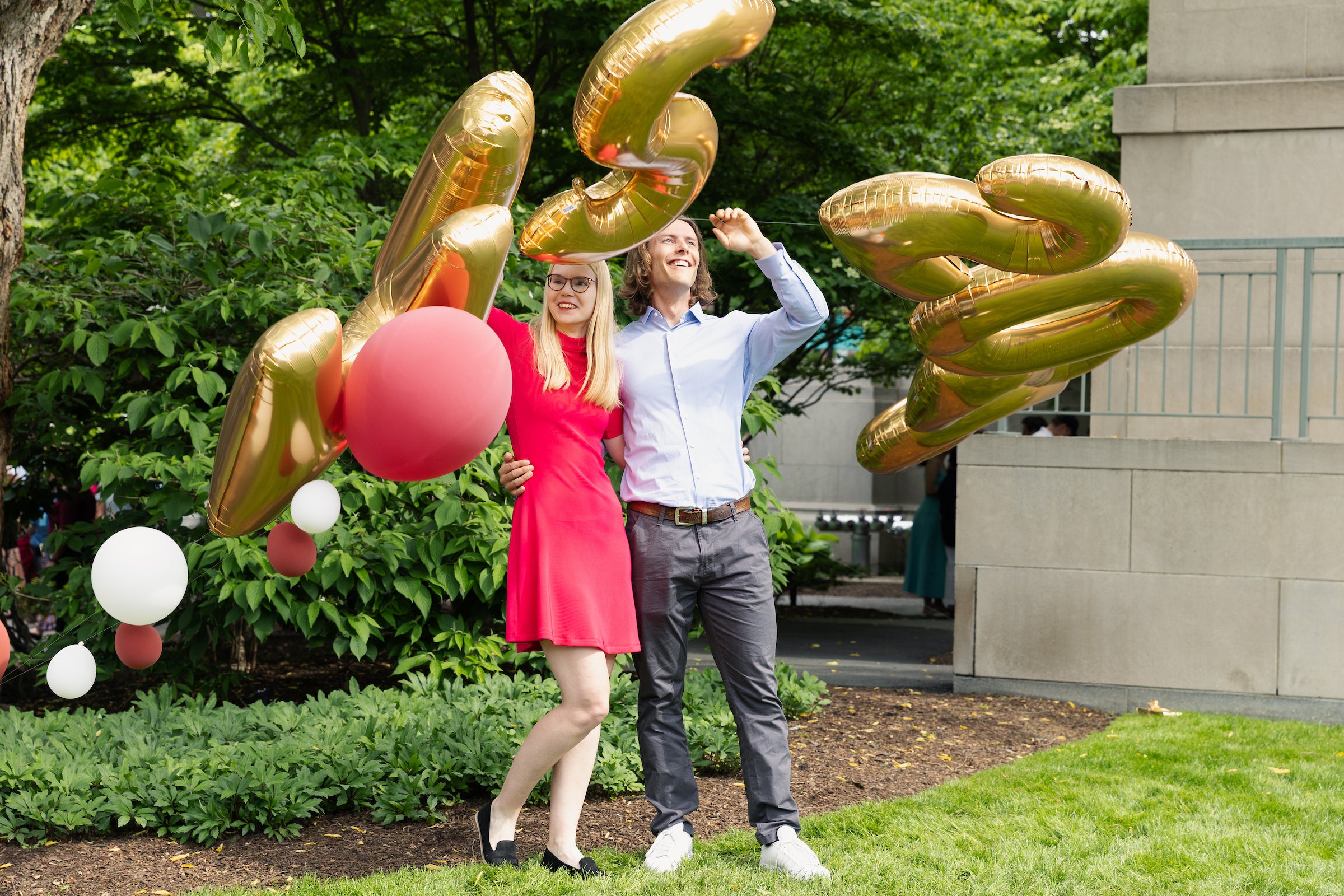 Two people stand outdoors and pose for a photo with balloons that are blowing in the wind.