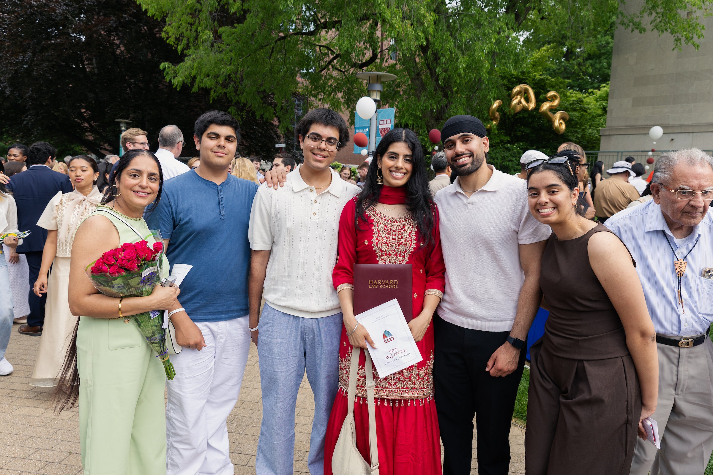 A group of six people pose together outdoors at a Harvard graduation event, with one person holding a program and another holding a bouquet of red roses.