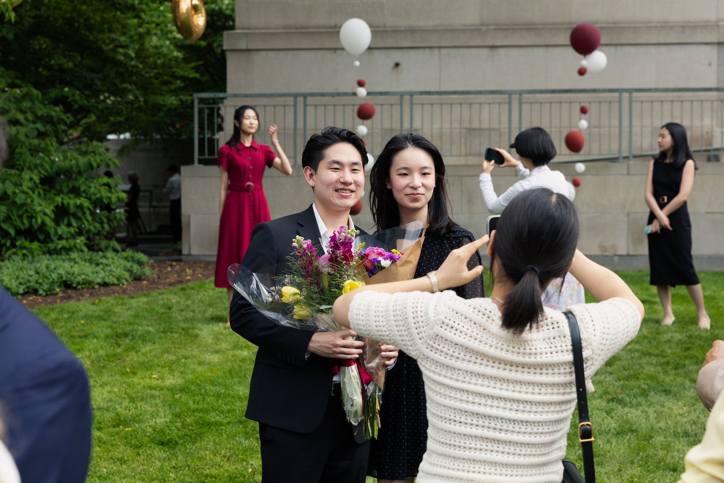 Two people smile for a photo outdoors while holding flowers, surrounded by attendees and festive balloons.