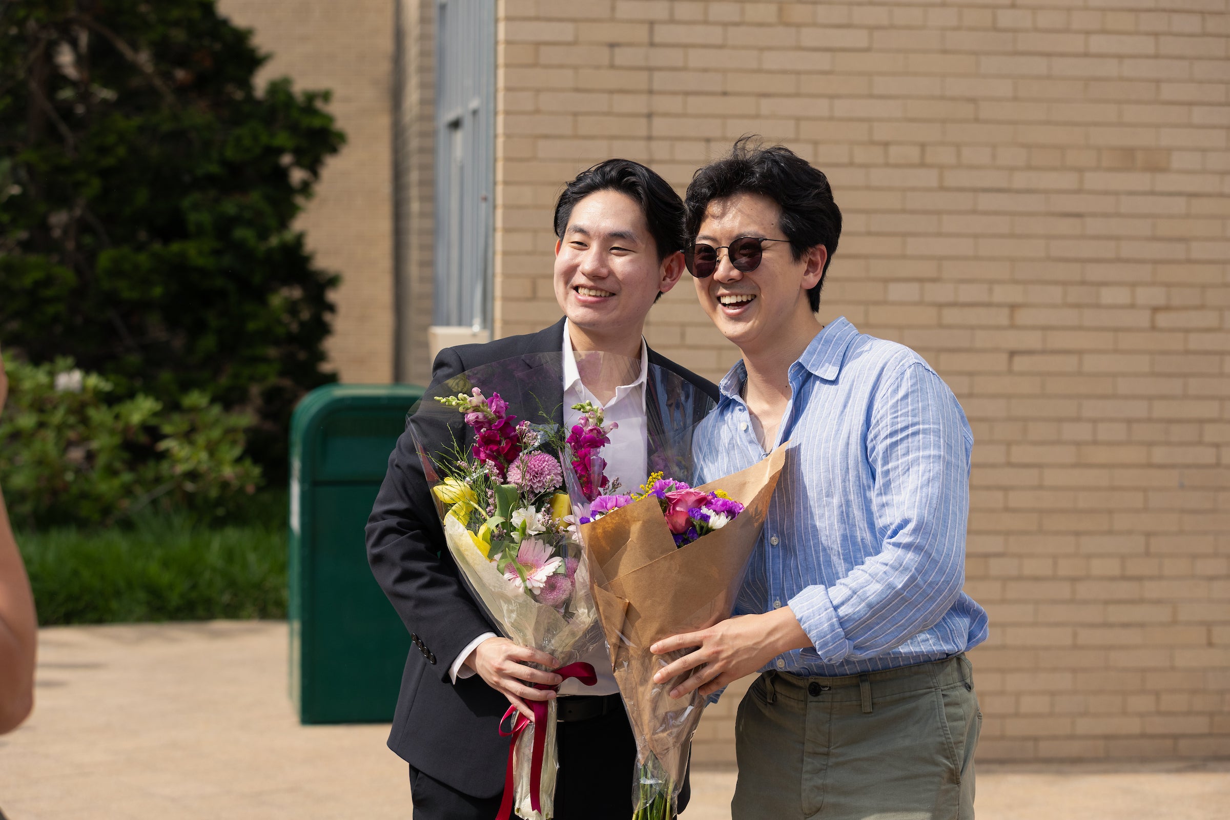 Two individuals stand outdoors, smiling and holding colorful flower bouquets, with a brick building and greenery in the background.