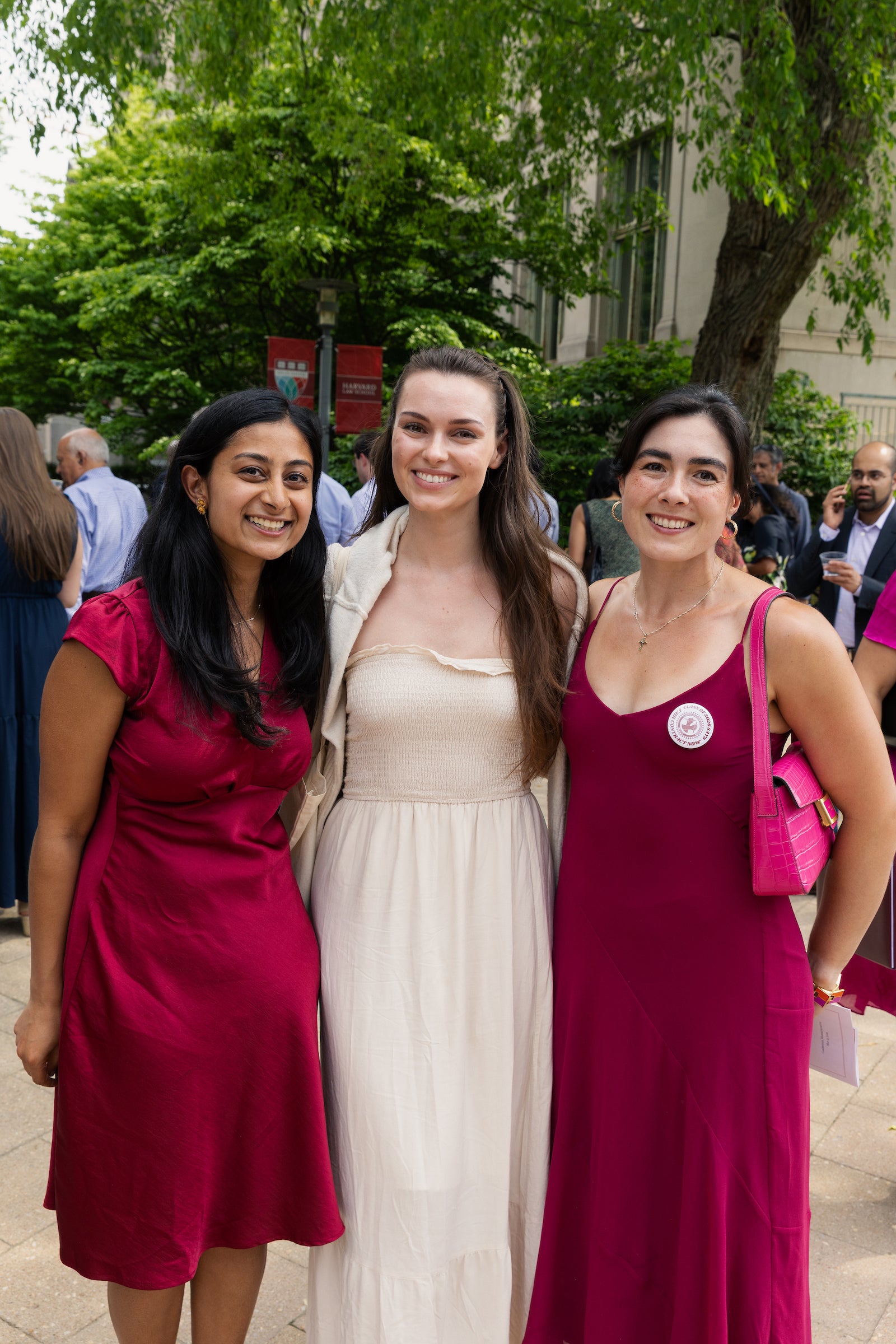 Three individuals wearing dresses pose together outdoors at an event, with trees and a building in the background.