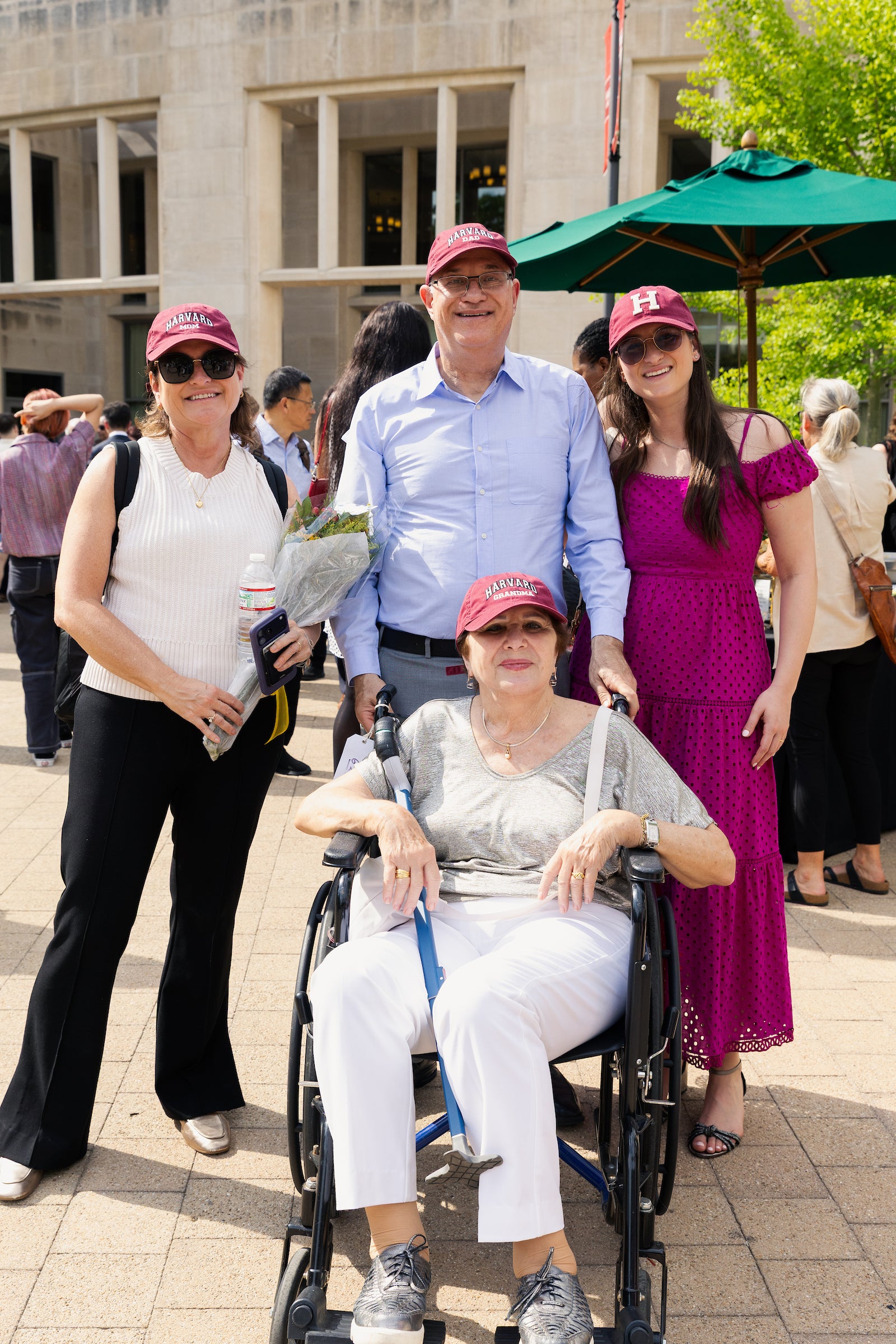 A group of four people, including one in a wheelchair, pose together outdoors wearing Harvard-themed caps in front of a stone building.