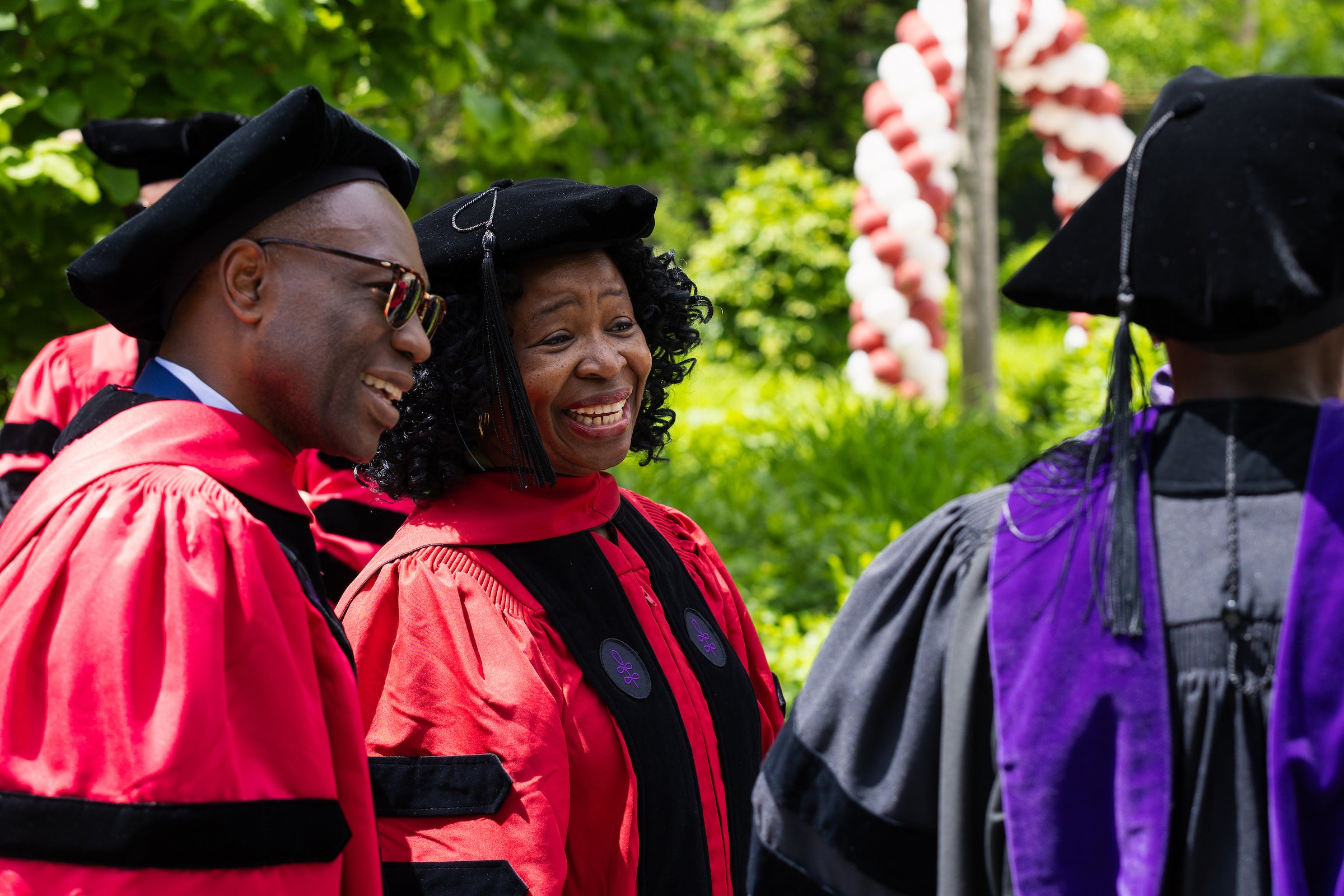 Harvard Law professors in academic regalia stand outdoors under trees, engaged in conversation.