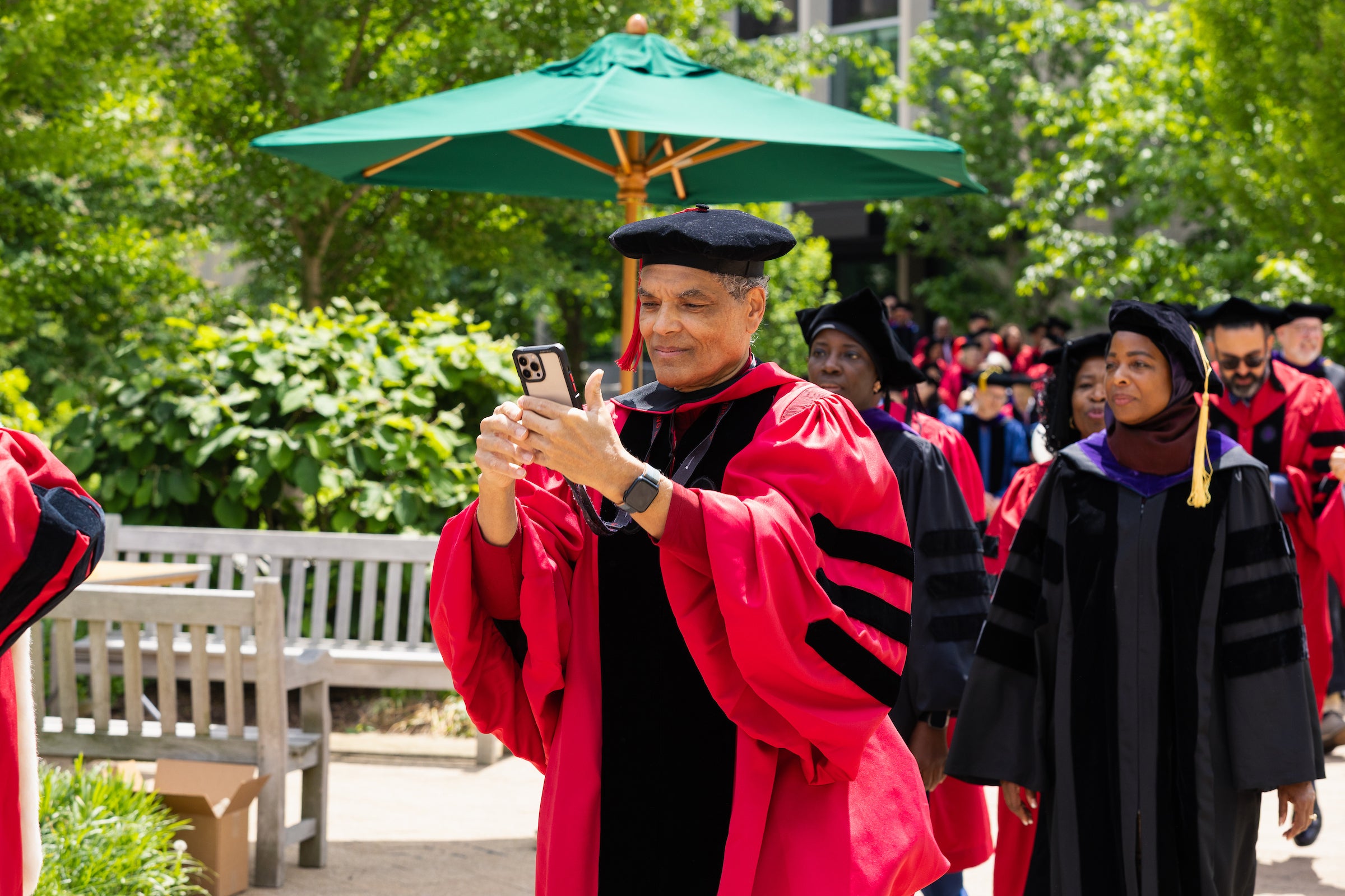 A Harvard Law professor in academic regalia captures a moment on their phone while walking outdoors in a line of other professors.