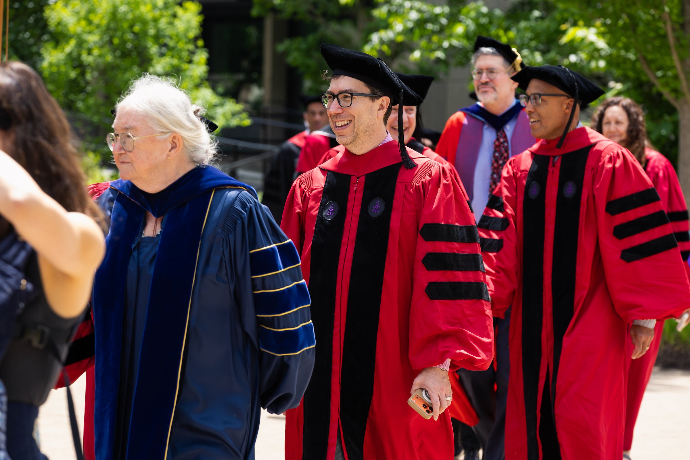A group of Harvard Law professors in academic regalia walk outdoors during a commencement ceremony under bright sunlight.