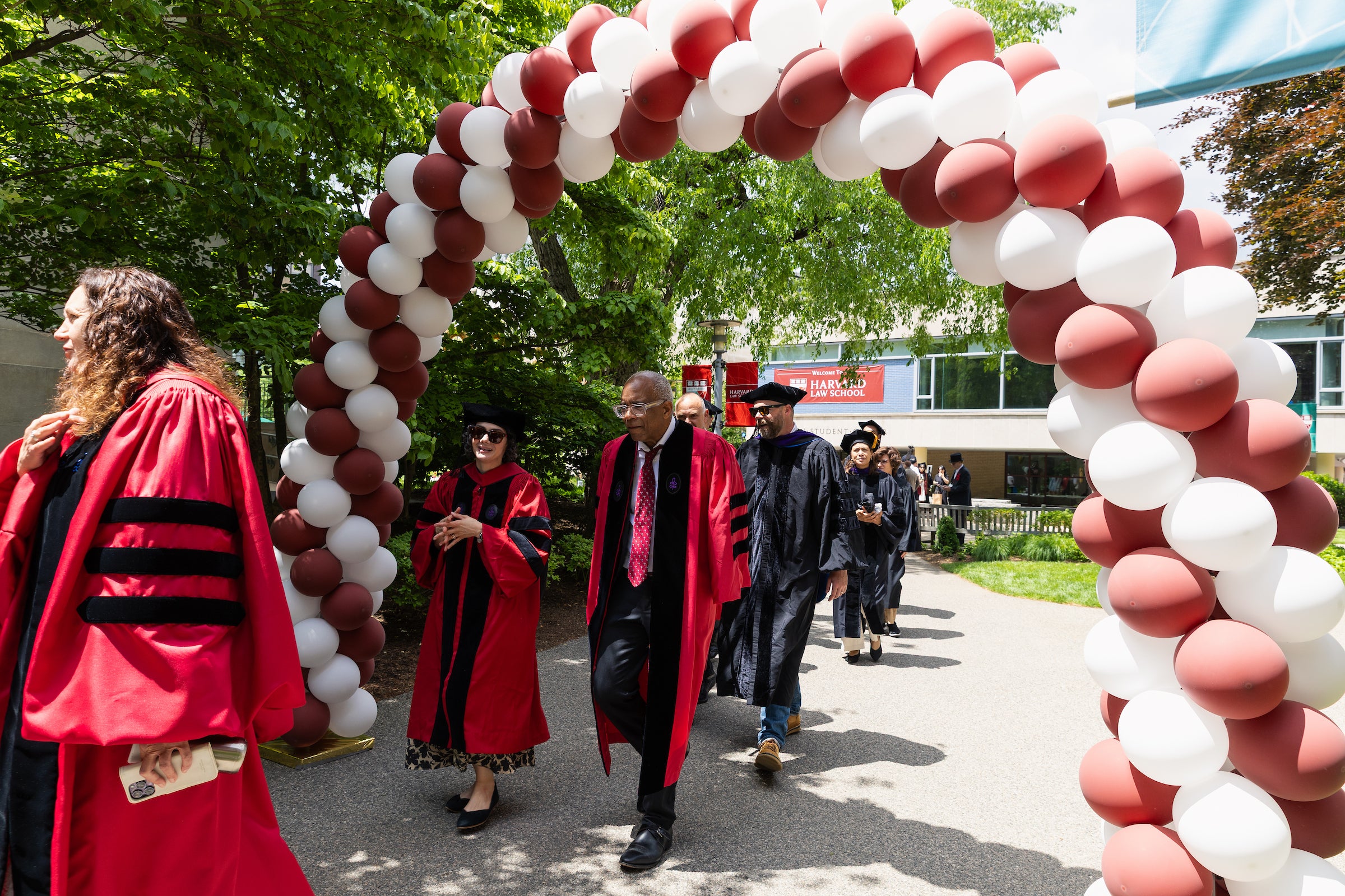 Harvard Law professors in academic regalia walk under a balloon arch during a procession.