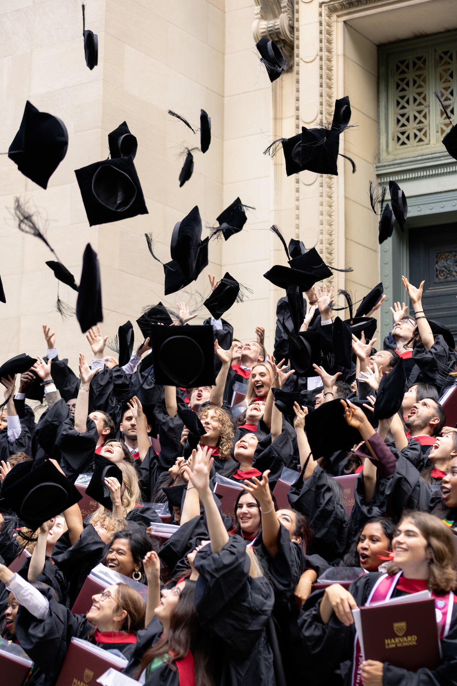 A crowd of graduates in black gowns toss their caps into the air outside a stone building.