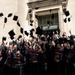 A crowd of graduates in black gowns toss their caps into the air outside a stone building.