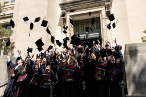 A crowd of graduates in black gowns toss their caps into the air outside a stone building.