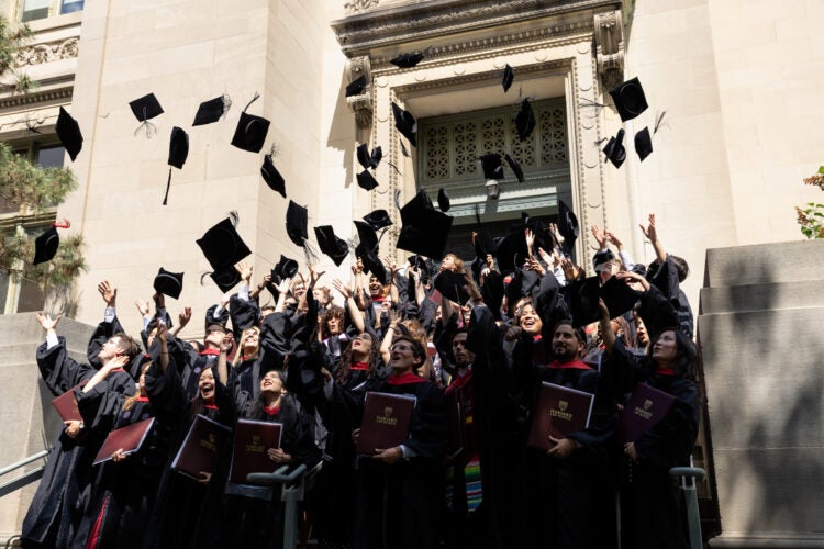 A crowd of graduates in black gowns toss their caps into the air outside a stone building.
