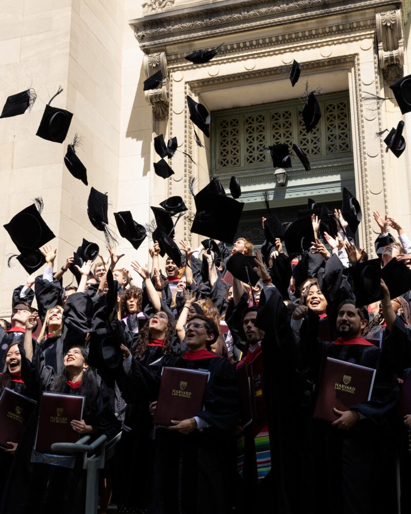 A crowd of graduates in black gowns toss their caps into the air outside a stone building.