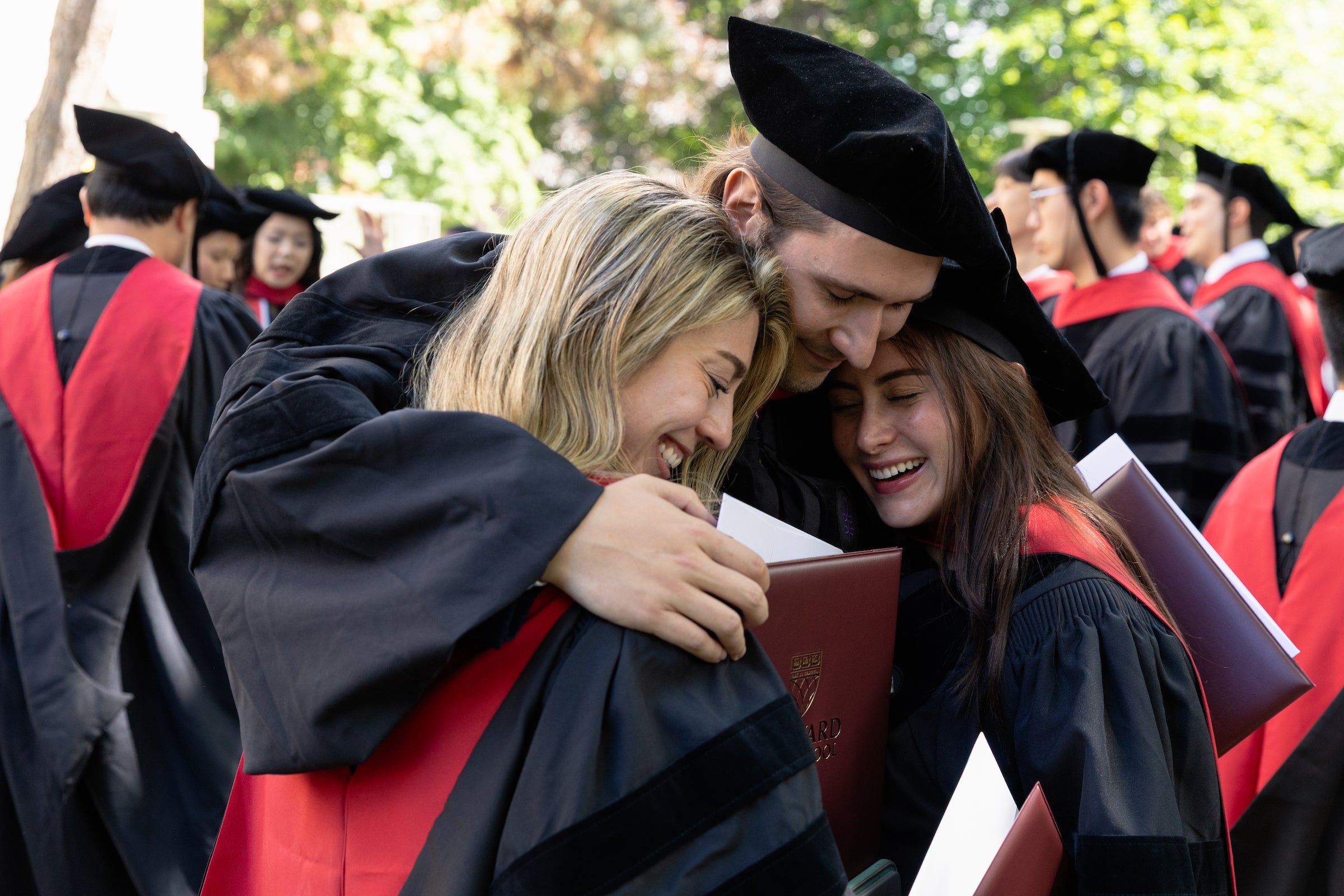 Three graduates in academic regalia embrace outdoors, holding diplomas.