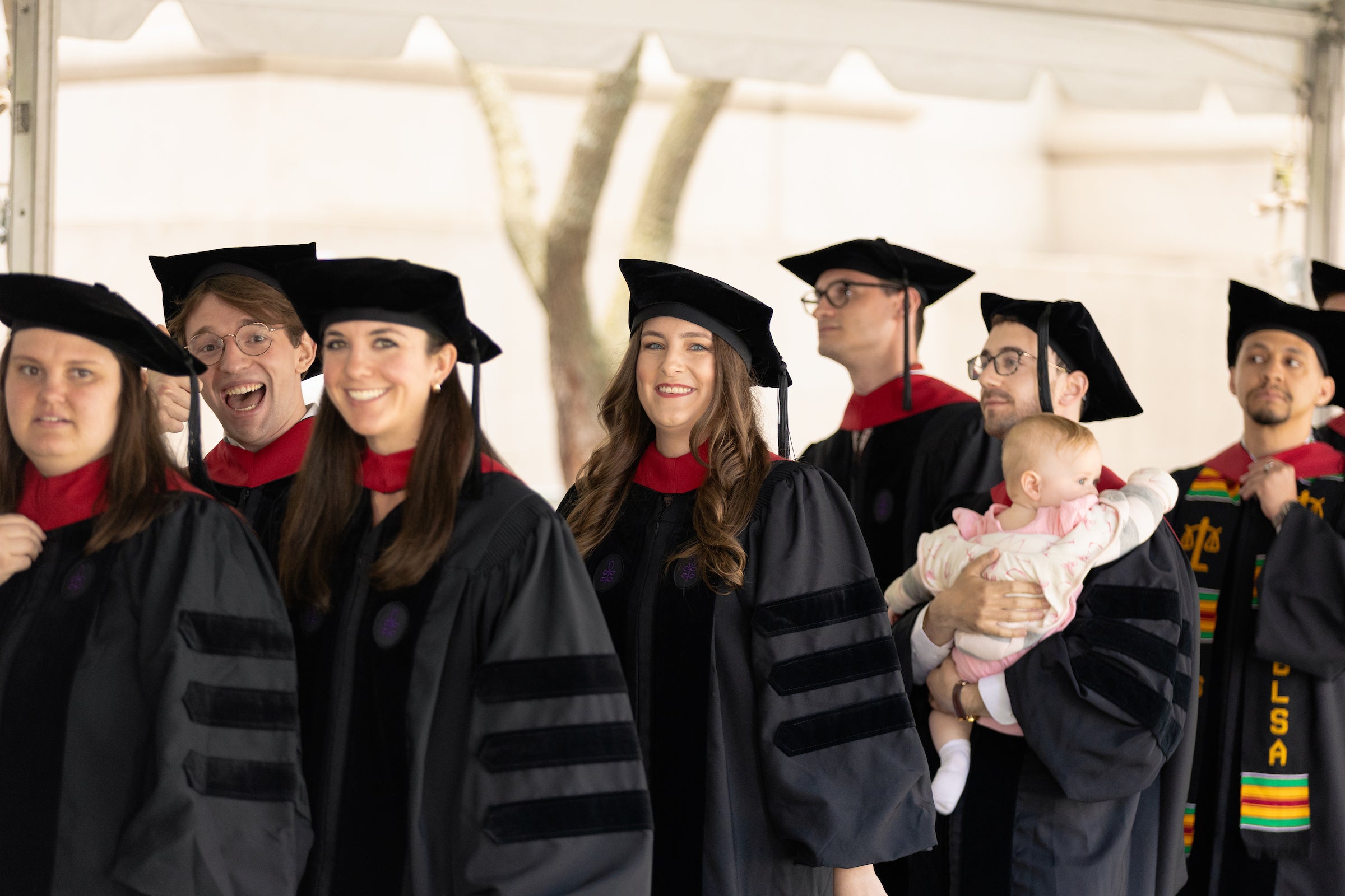 A group of graduates in black academic regalia and caps wait in line under a tent, smiling and waiting to accept their diplomas, with one person holding a baby.