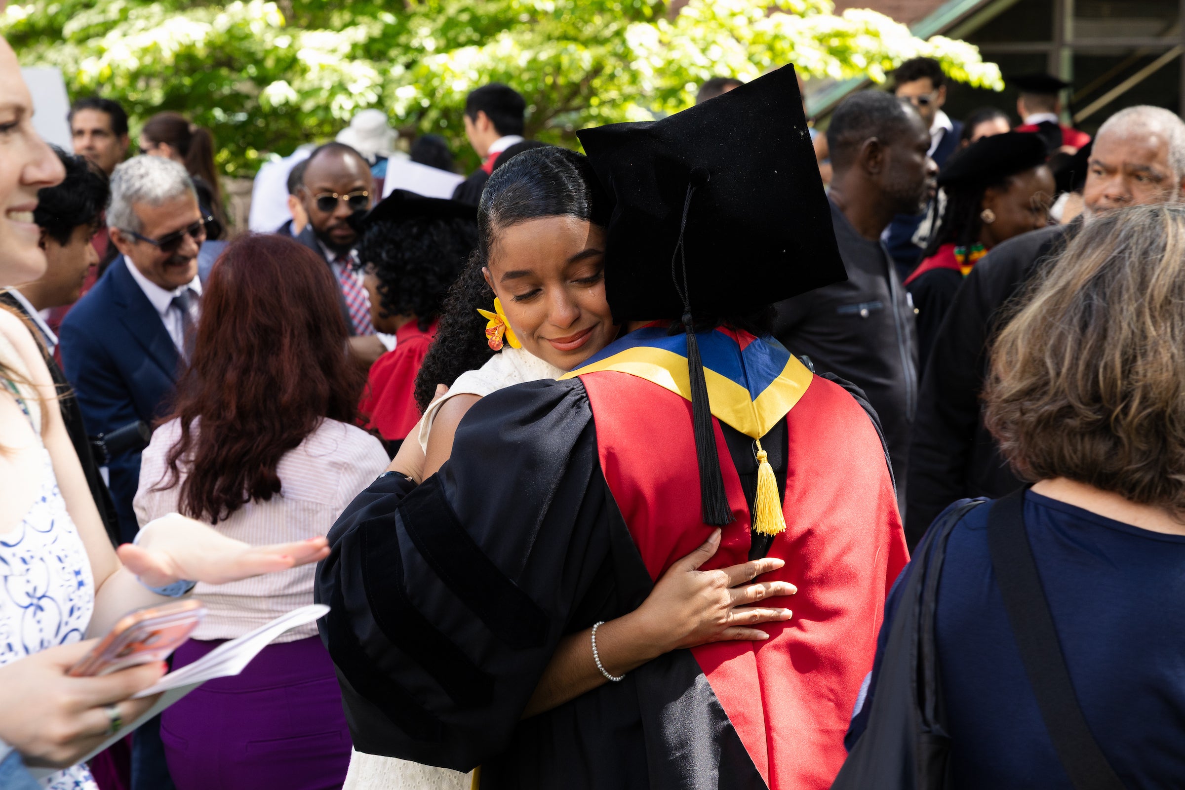 A graduate in a black and red academic gown embraces another person at an outdoor ceremony, surrounded by attendees.