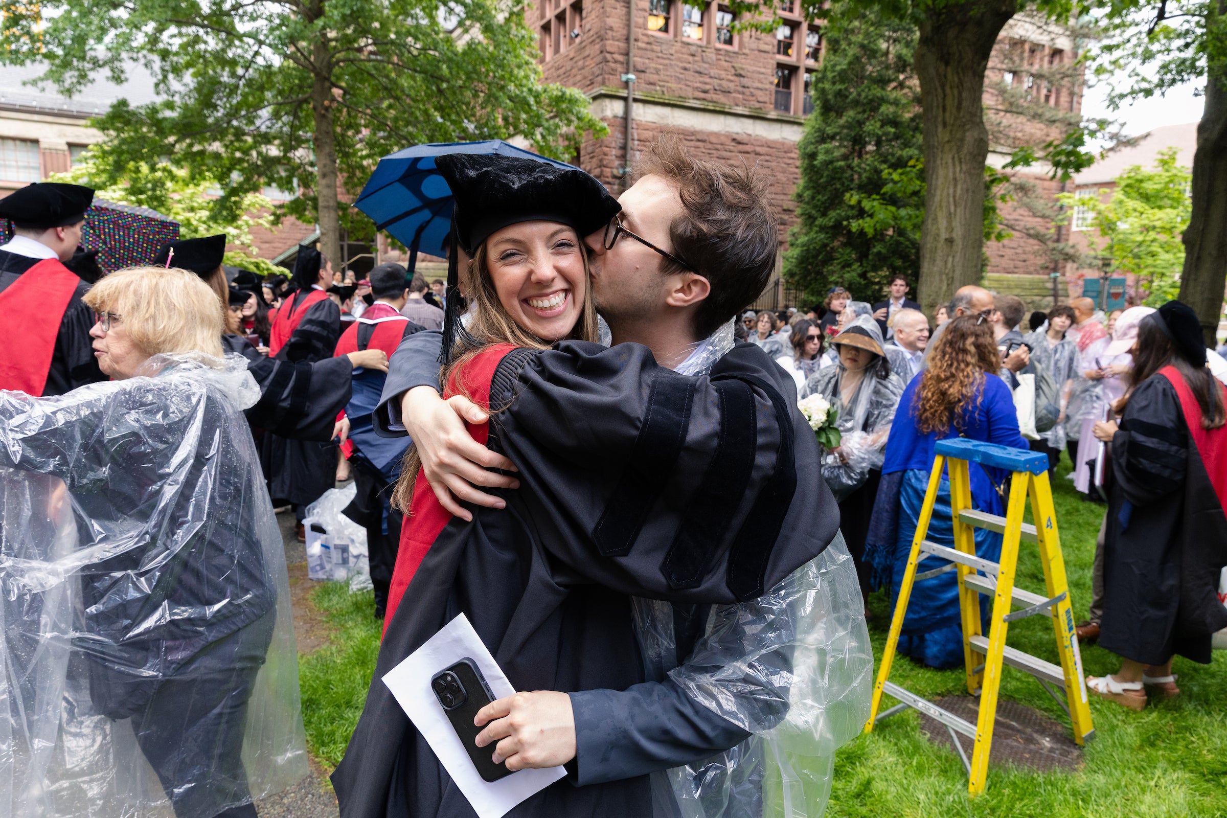 A graduate smiles for the camera as her partner kisses her cheek.