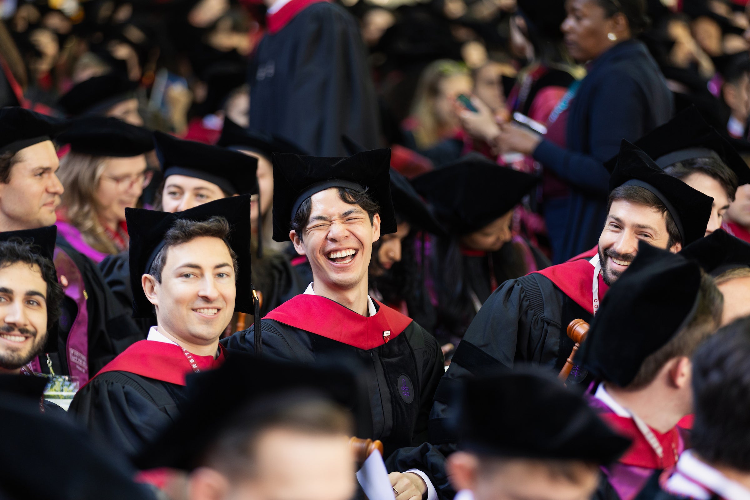 Graduates in black caps and gowns with red stoles laugh joyfully during a commencement ceremony.