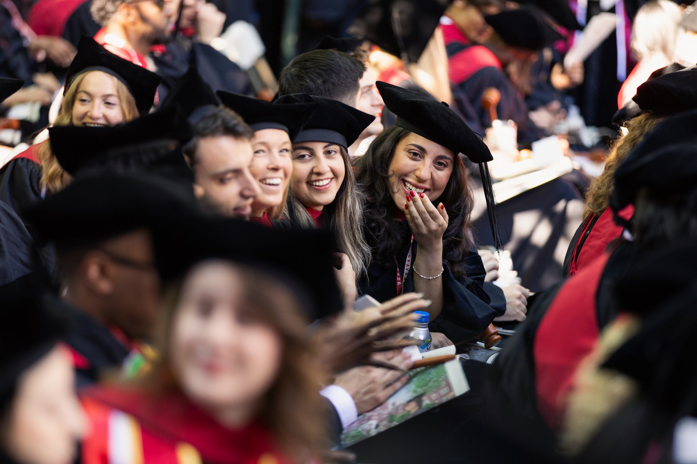 Graduates in black caps and gowns with red stoles laugh in their seats during a commencement ceremony.