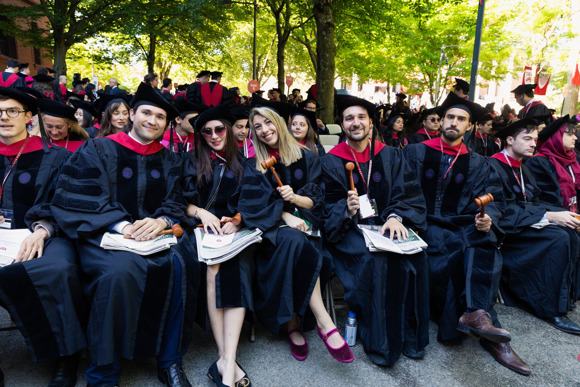 Graduates in black and red academic regalia sit outdoors, some holding wooden gavels and diplomas, surrounded by trees and fellow attendees.
