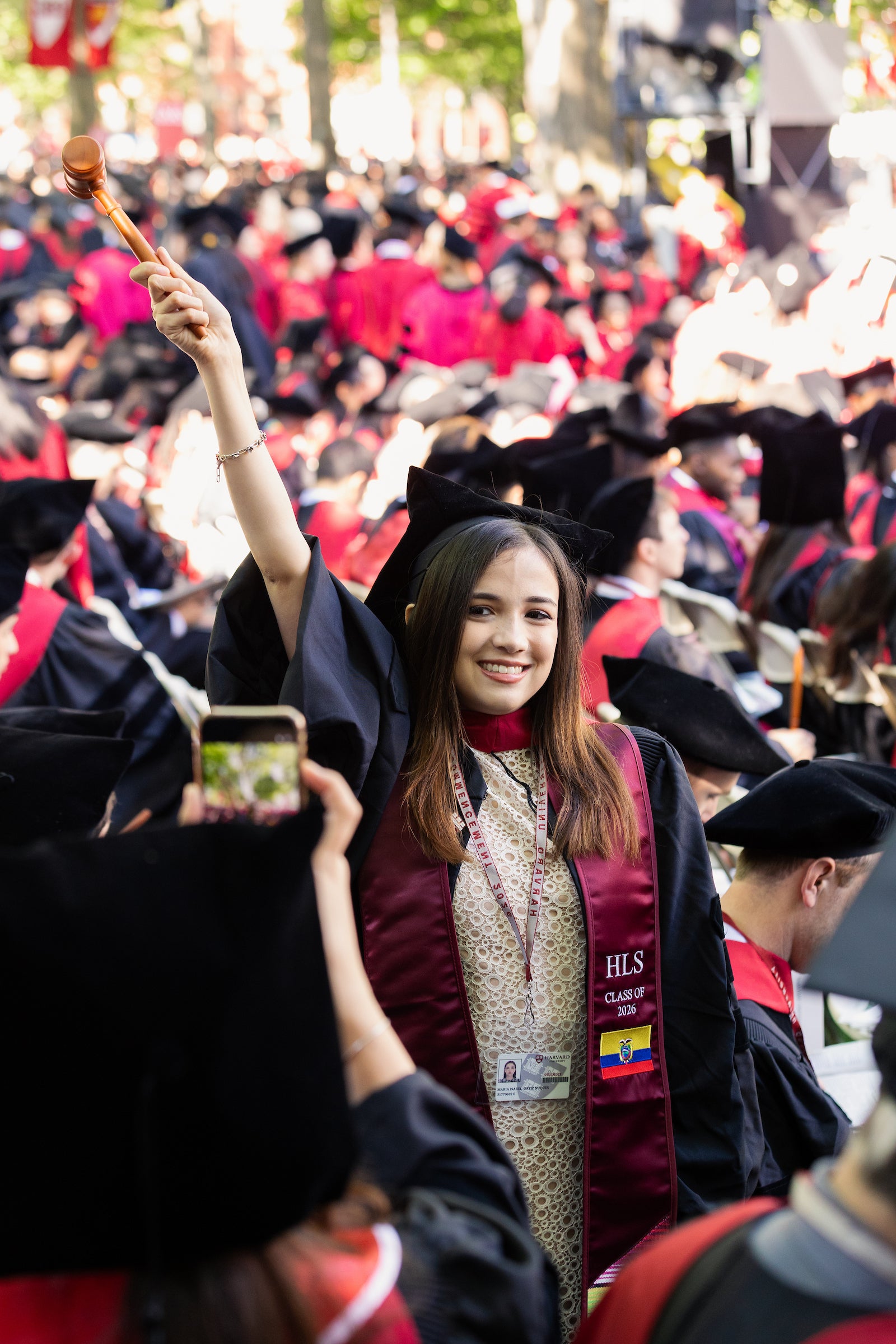 A graduate in a maroon and black academic regalia poses for a photo by raising a wooden gavel in celebration during a large outdoor commencement ceremony.
