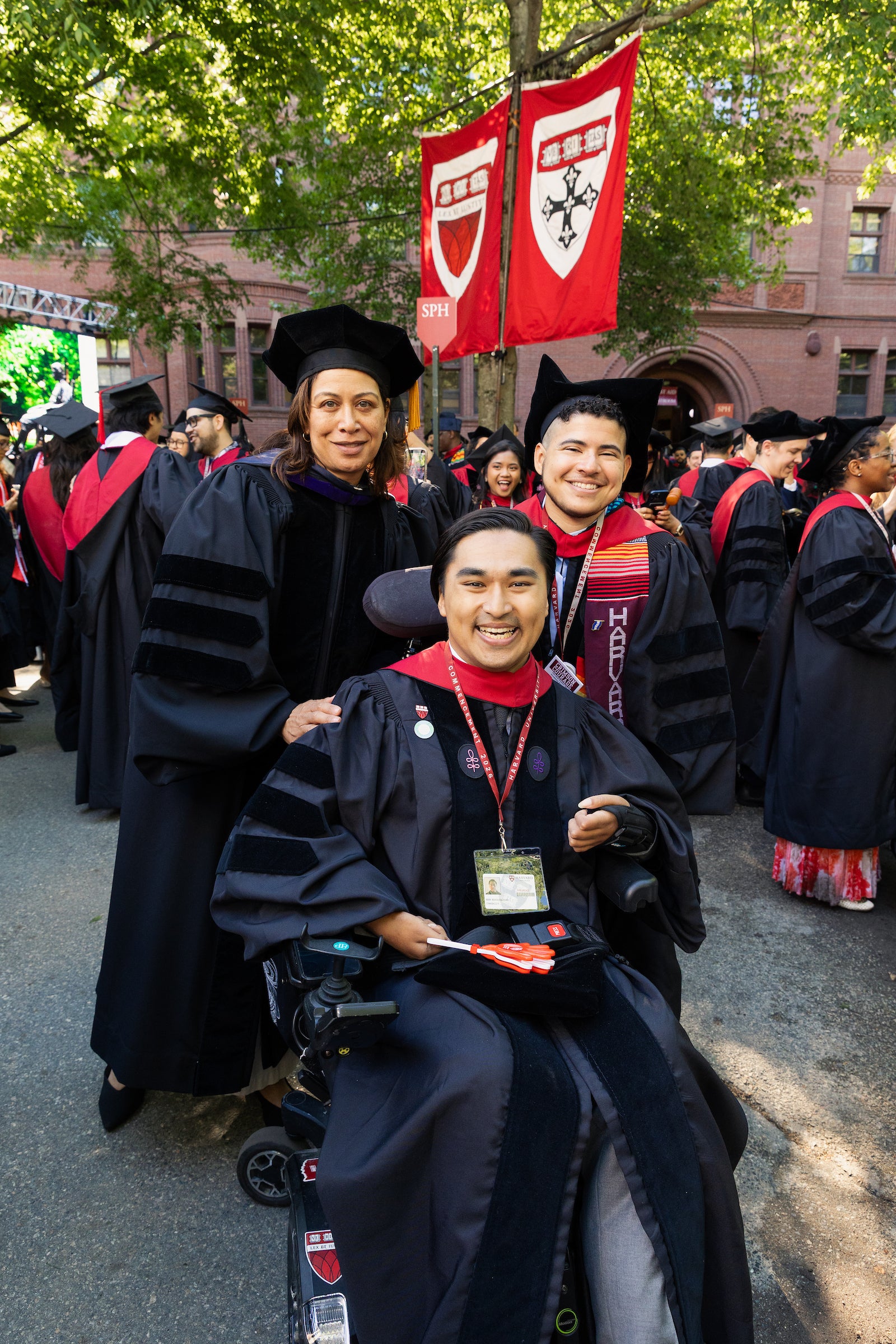 A graduate in a wheelchair smiles for a photo with two others in regalia at a commencement ceremony under trees with campus banners.
