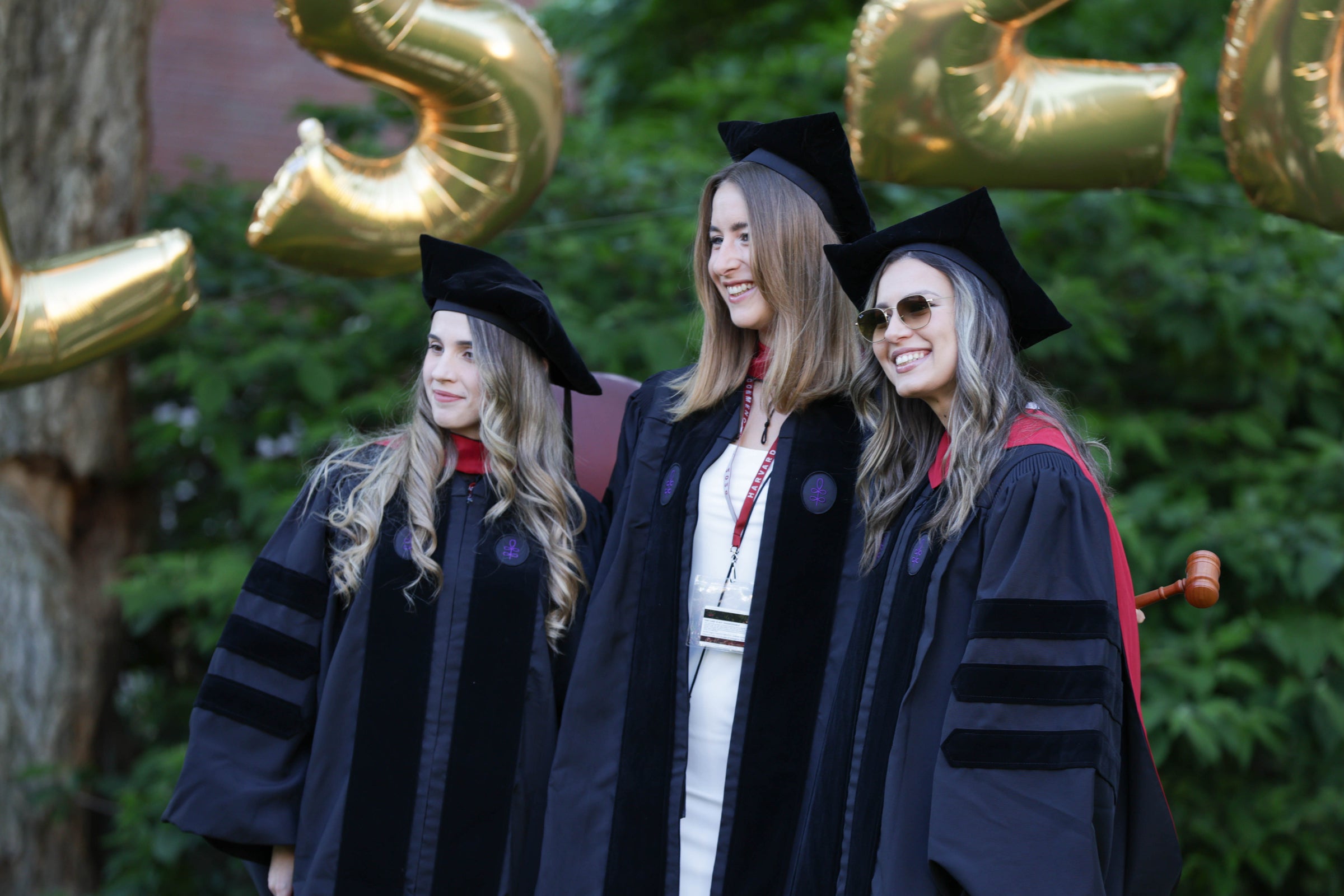 Three graduates in regalia smile for the camera outdoors, with large gold balloons visible in the background.