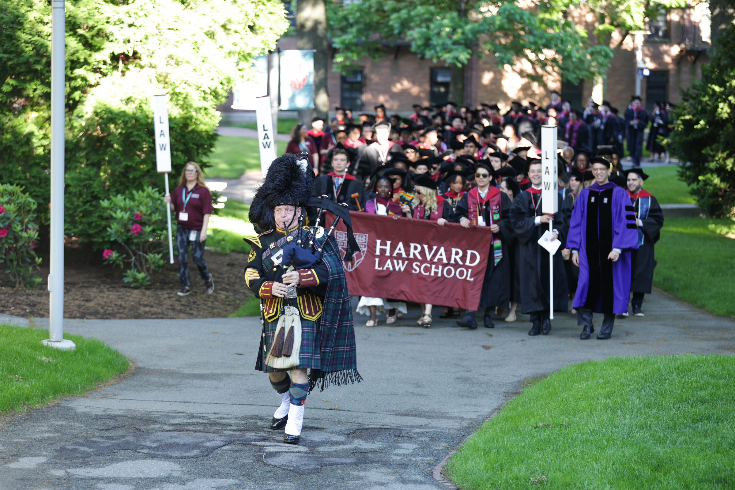 A bagpiper in traditional attire leads a procession of graduates carrying a Harvard Law School banner along a campus pathway.