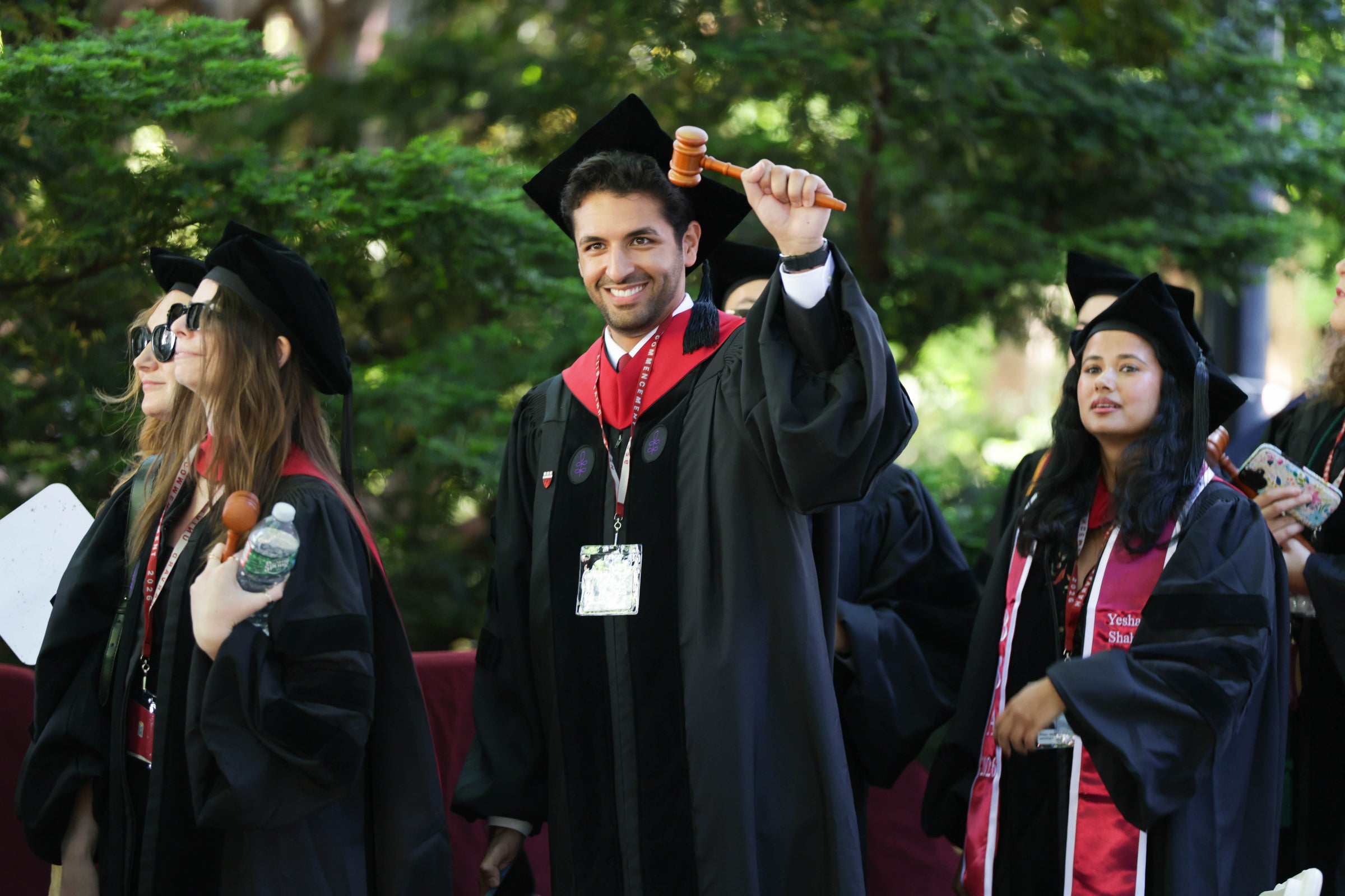 A graduate in regalia smiles while holding a wooden gavel, surrounded by other graduates outdoors.