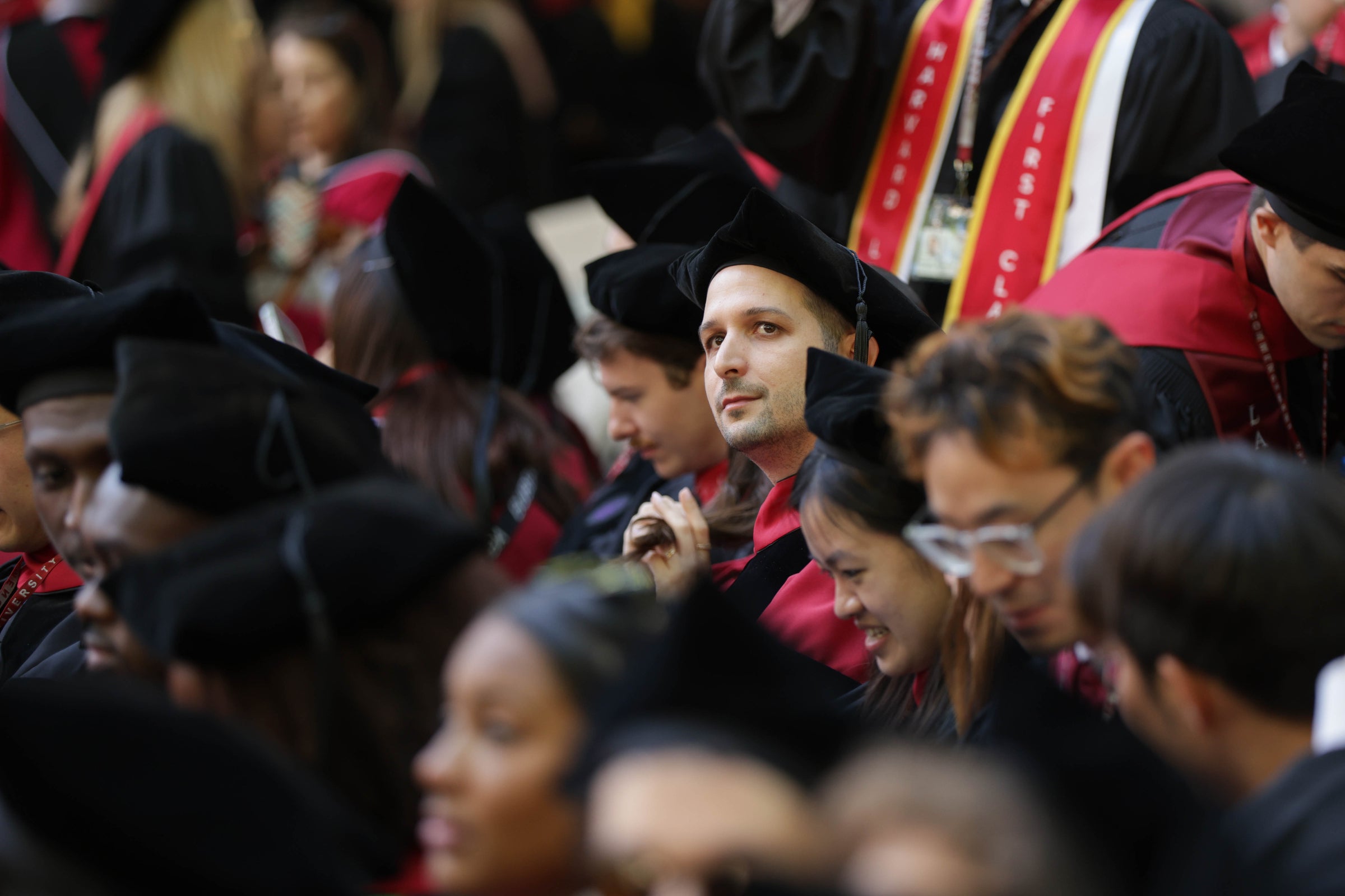A crowd of graduates in caps and gowns are seated for commencement, with one individual in a red and black gown looking forward attentively.