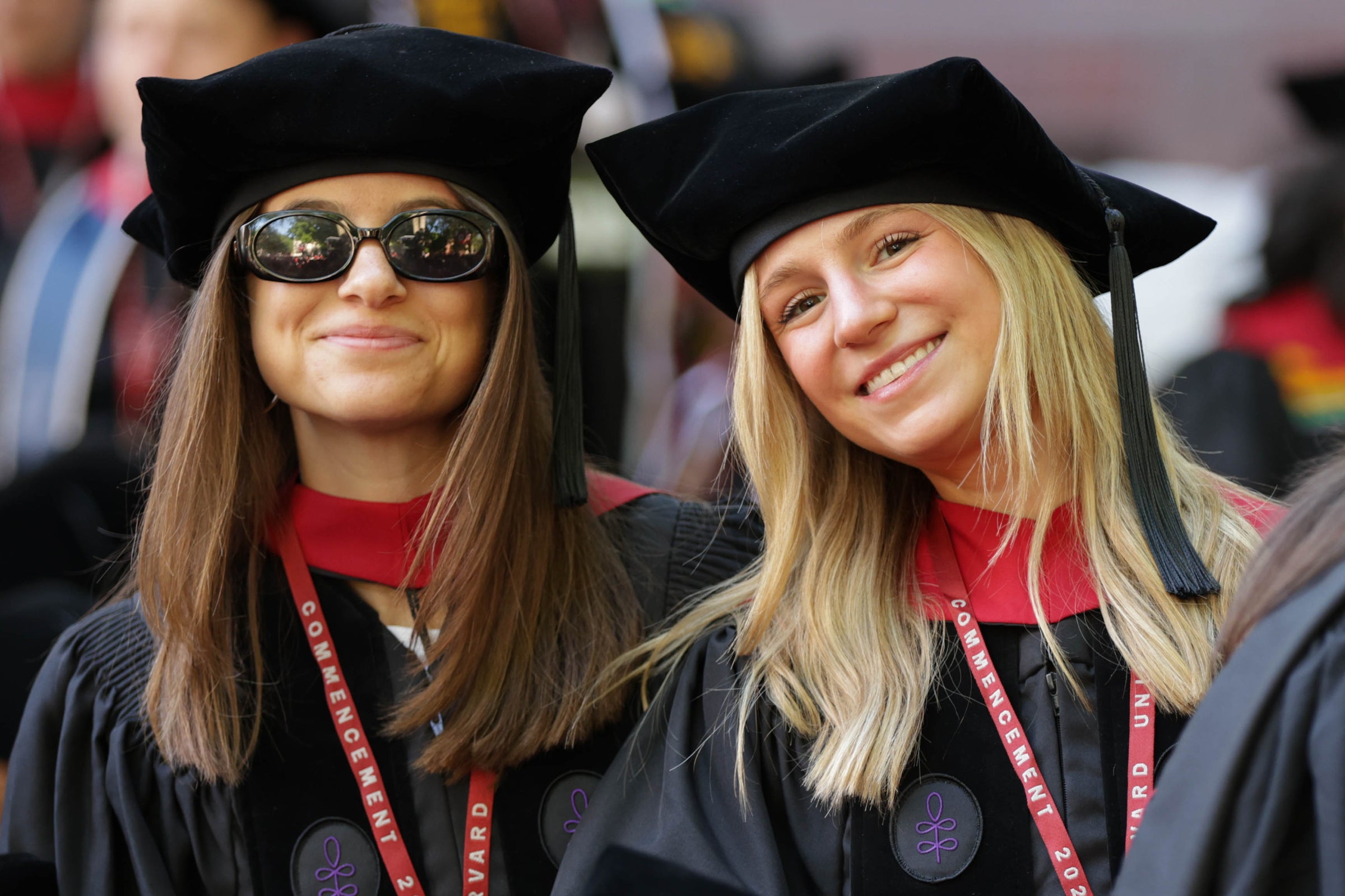 Two graduates in black caps and gowns with red lanyards smile at the camera during a Harvard commencement ceremony.