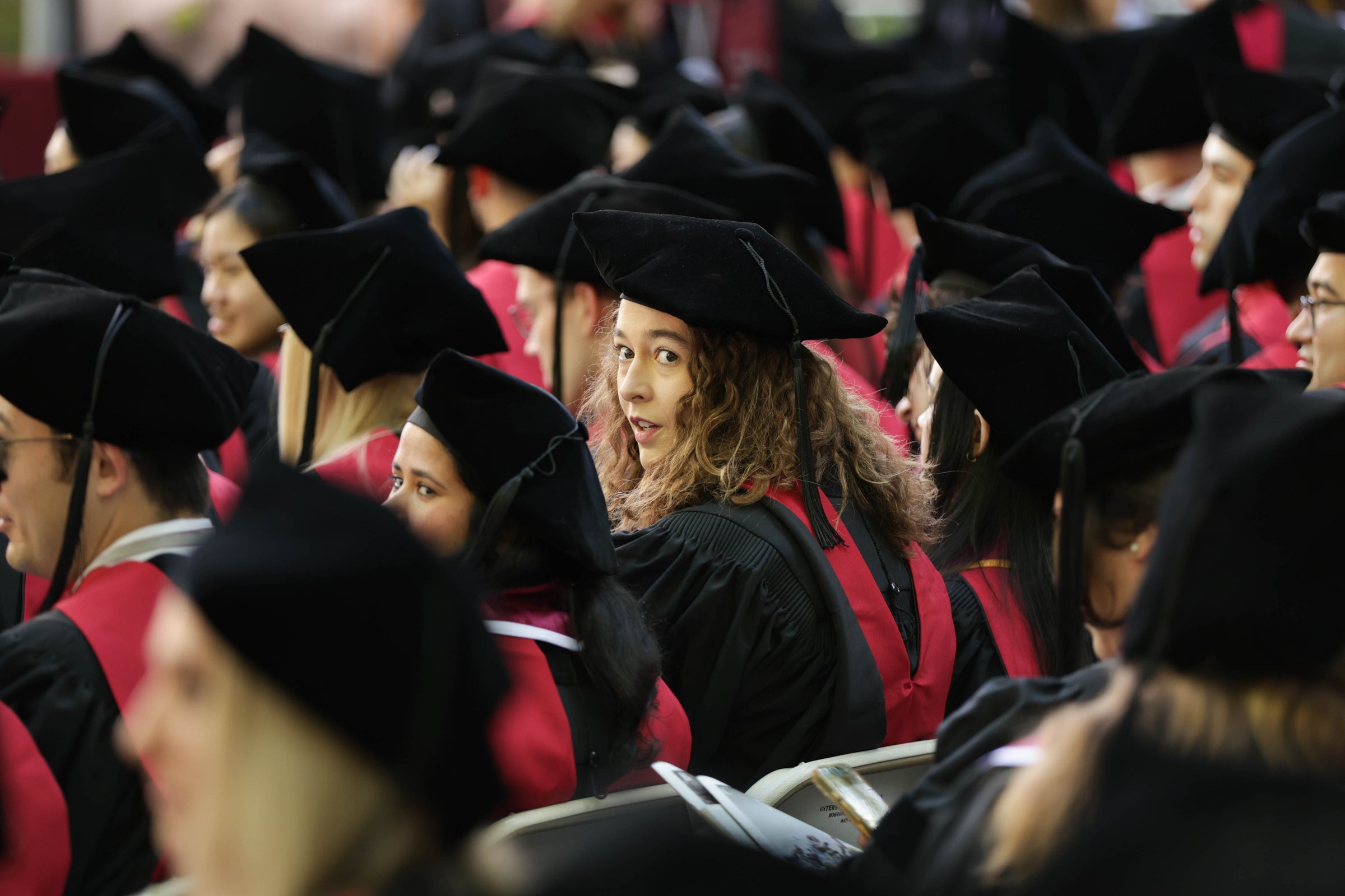 A graduate with curly hair looks over her shoulder amidst a crowd of peers in black caps and red-trimmed gowns during a commencement ceremony.