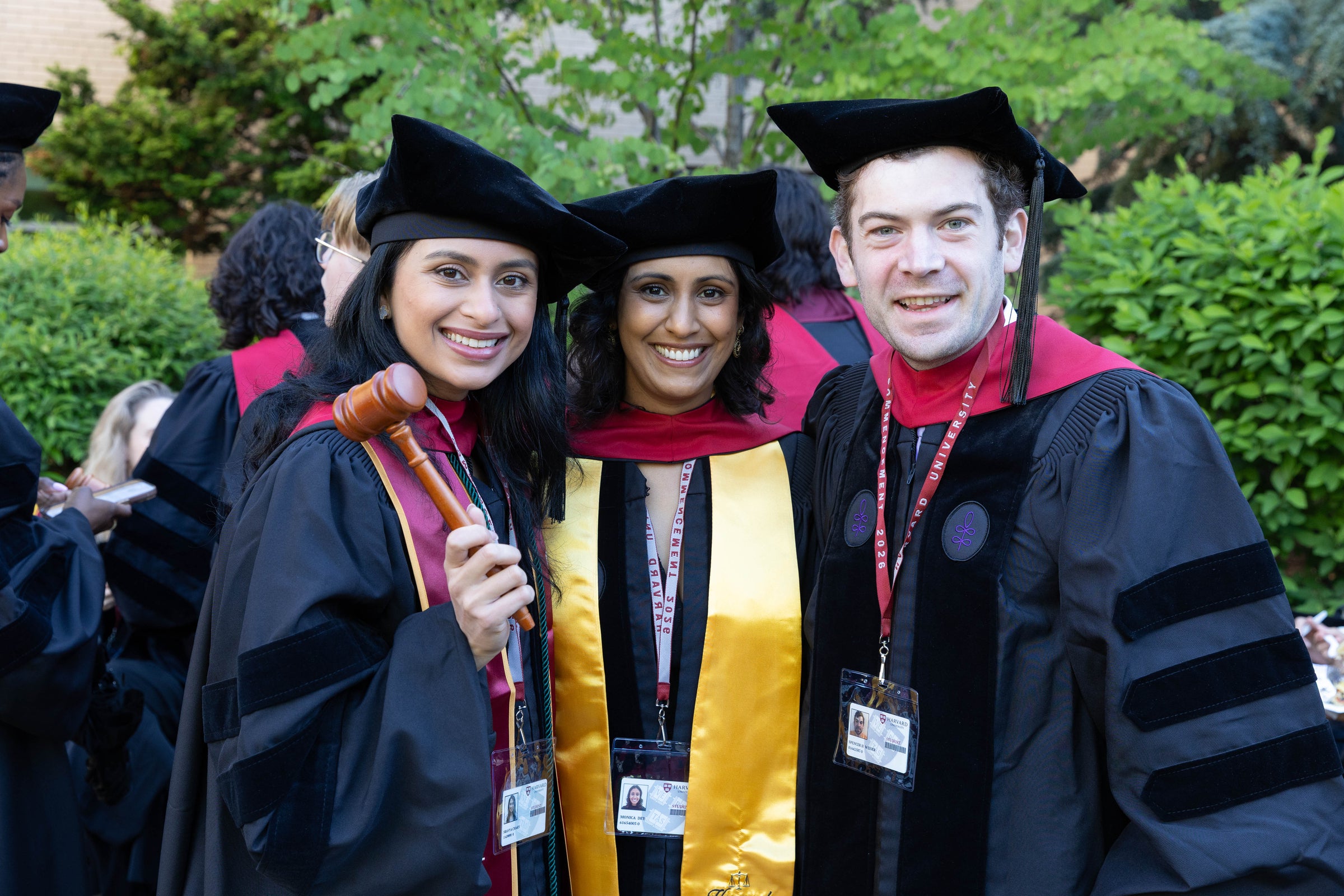 Three graduates in regalia smile for the camera, one holding a wooden gavel, at an outdoor ceremony.