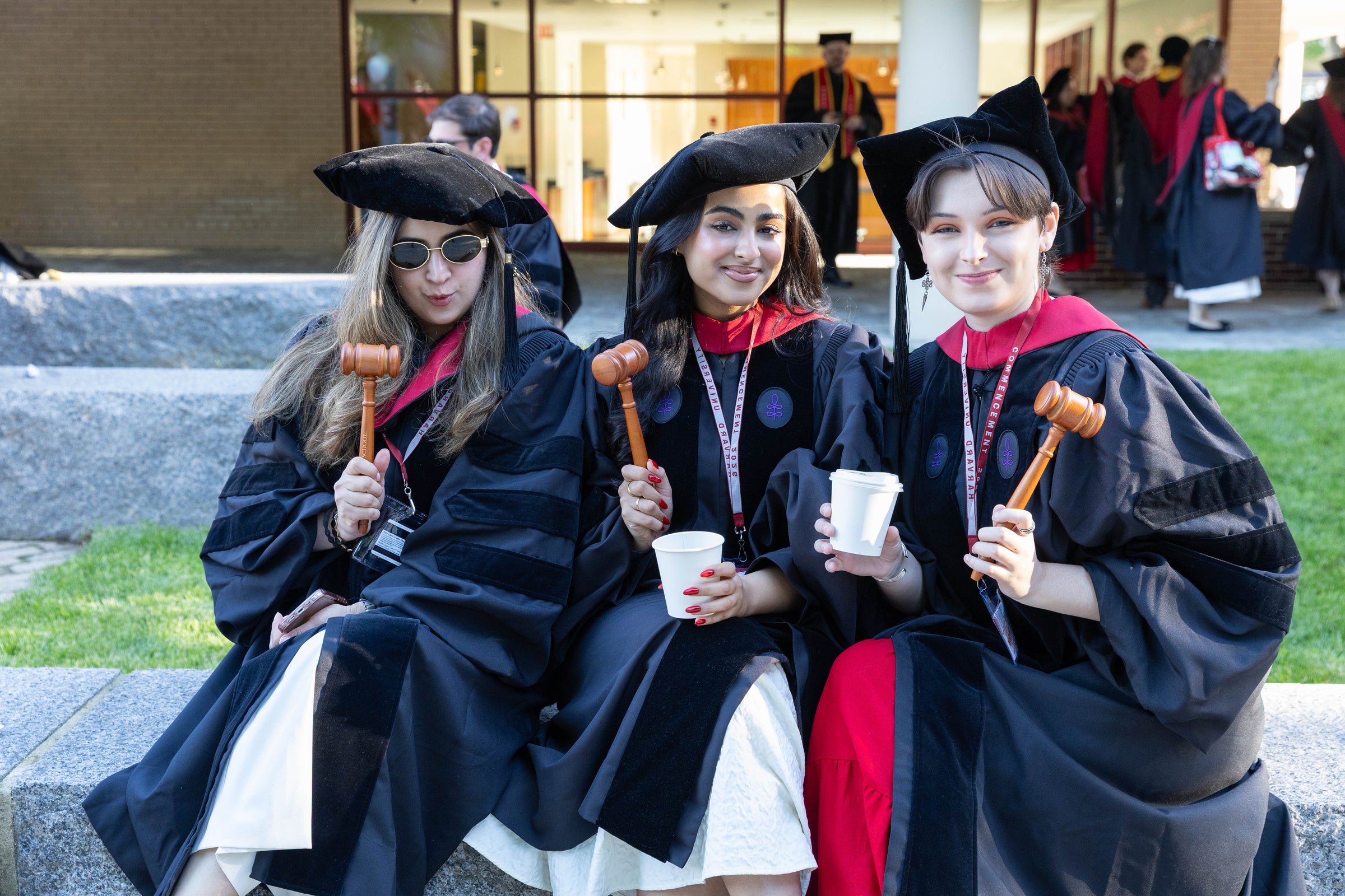 Three graduates in regalia sit outdoors, holding gavels and cups, smiling for the camera.