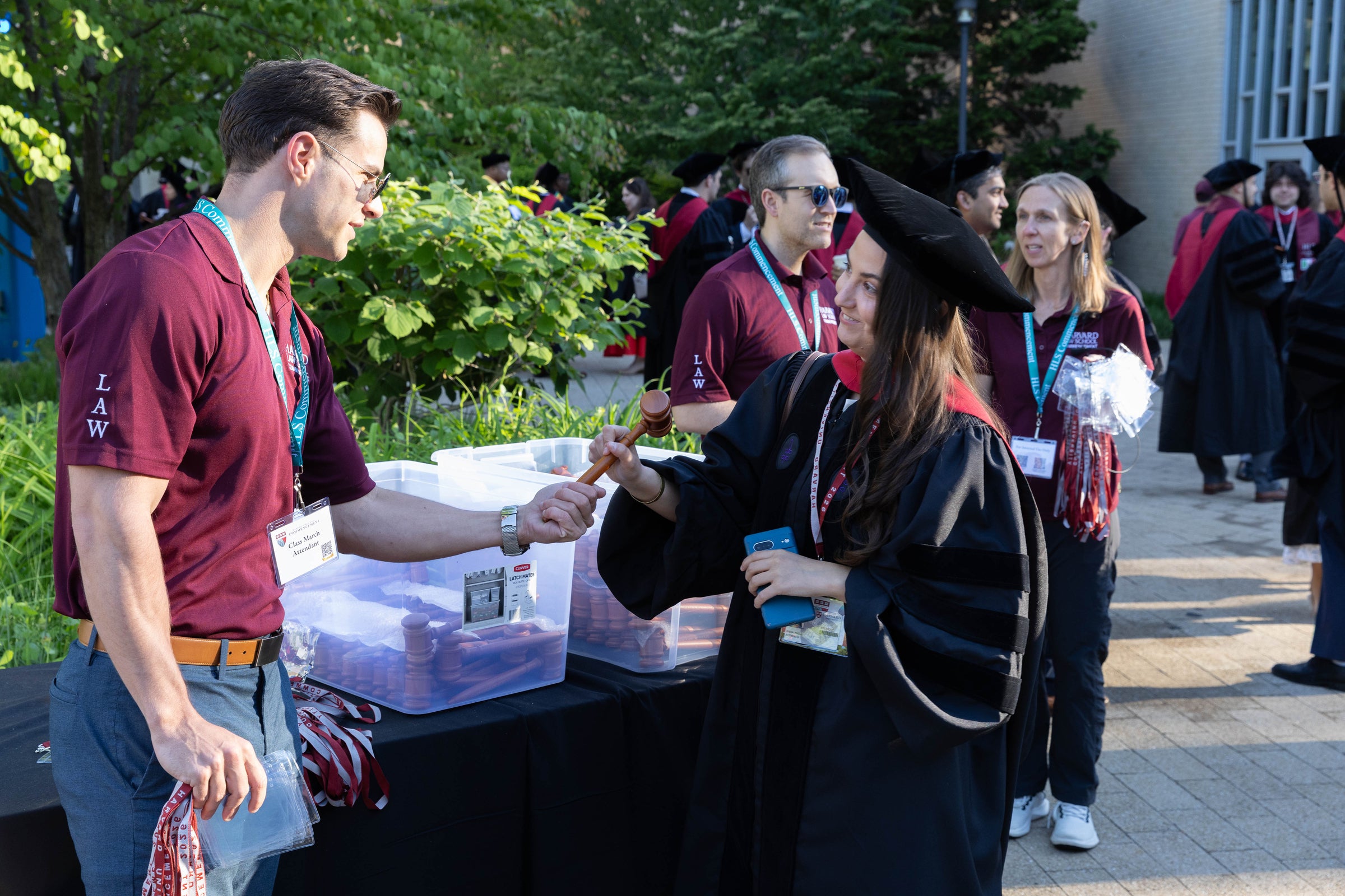 A graduate in regalia receives a wooden gavel from a volunteer at an outdoor event.