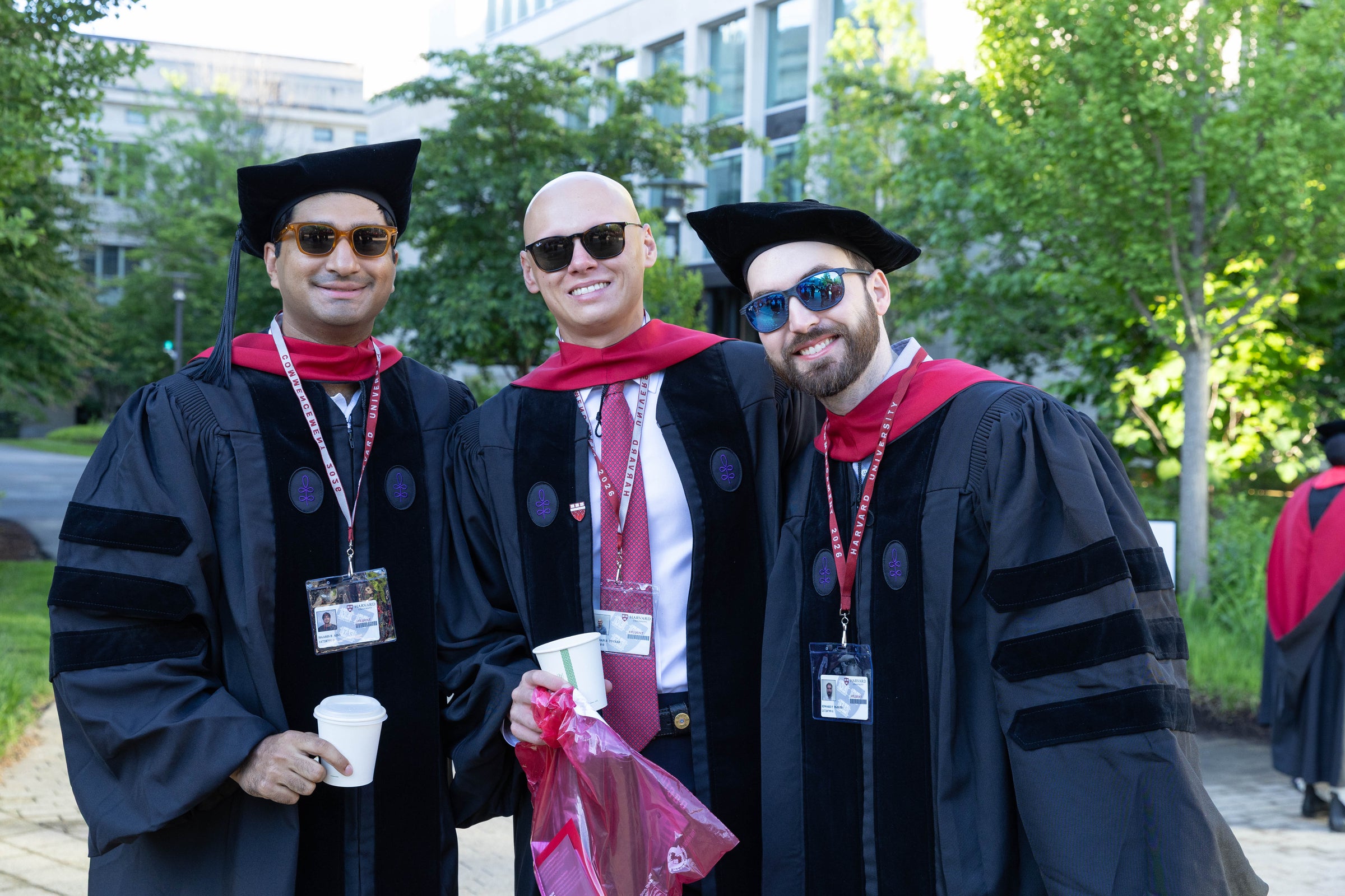 Three individuals in academic regalia pose outdoors, smiling while holding drinks and a plastic bag.