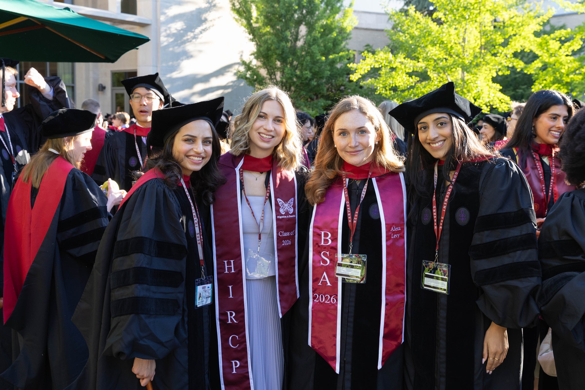 A group of graduates in black caps and gowns with maroon sashes stand together and smile for the camera in an outdoor courtyard.
