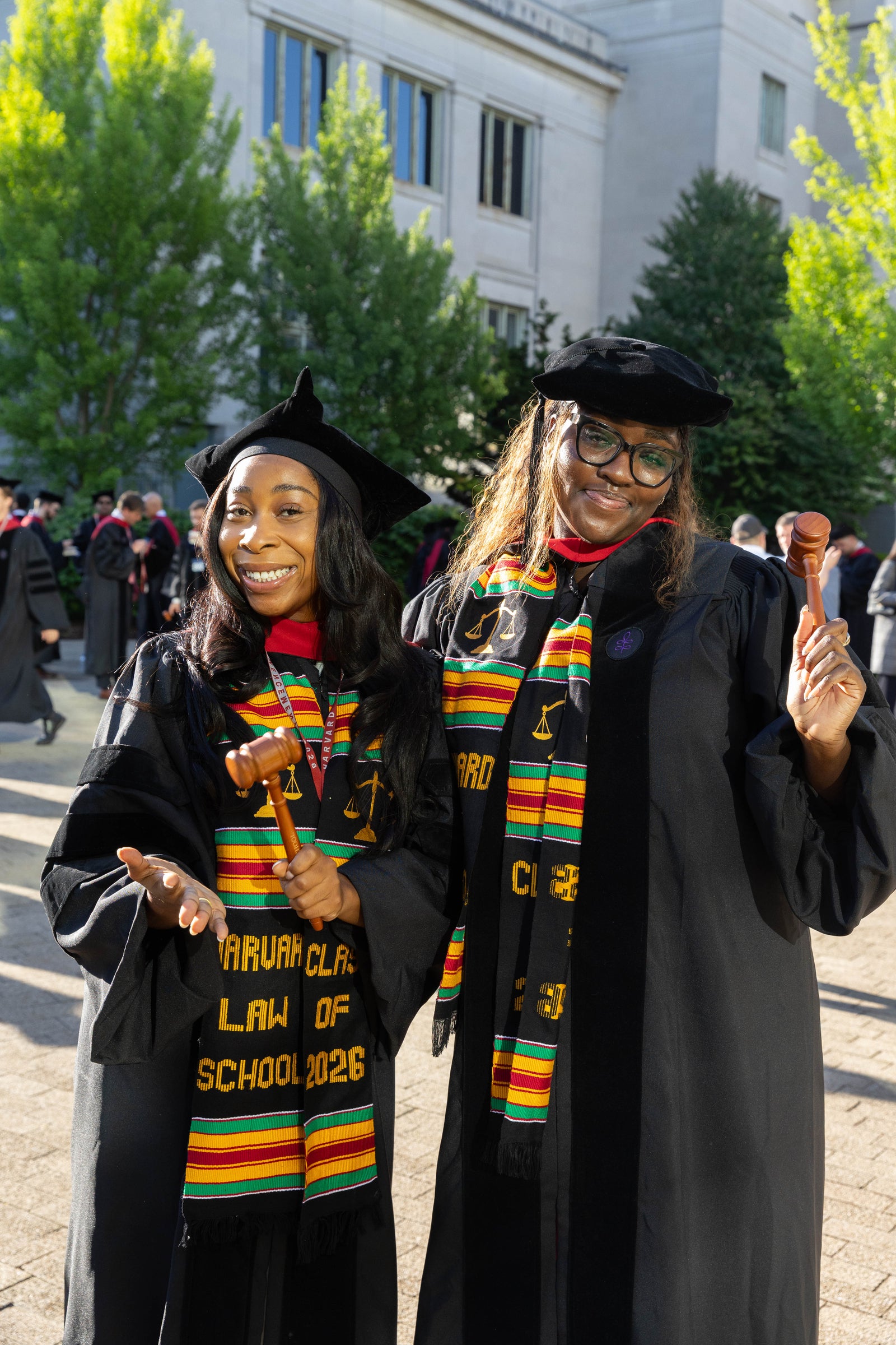Two graduates in regalia pose for a photo outdoors while holding up gavels.