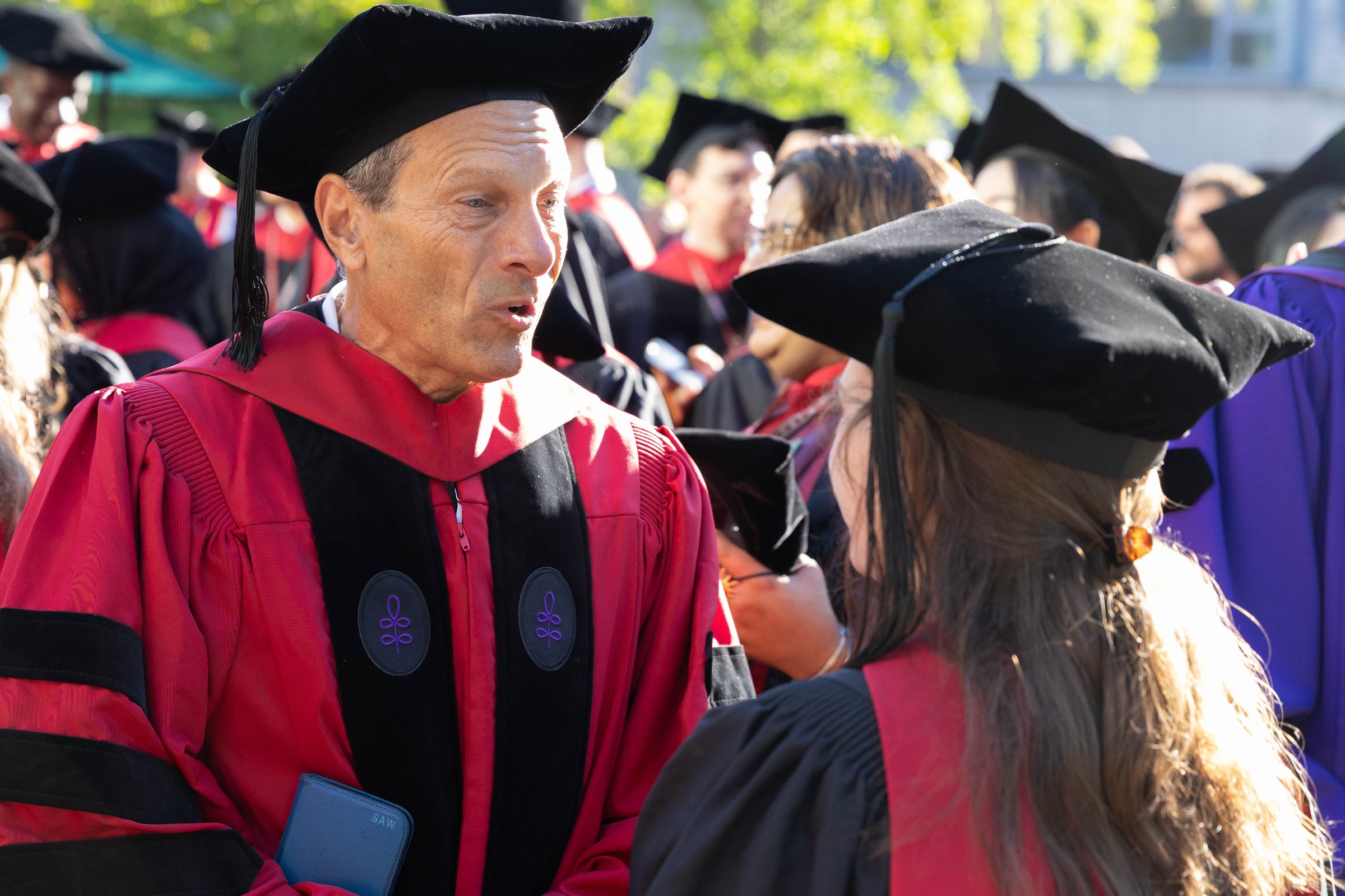 A faculty member in academic regalia speaks with a graduate at an outdoor event.