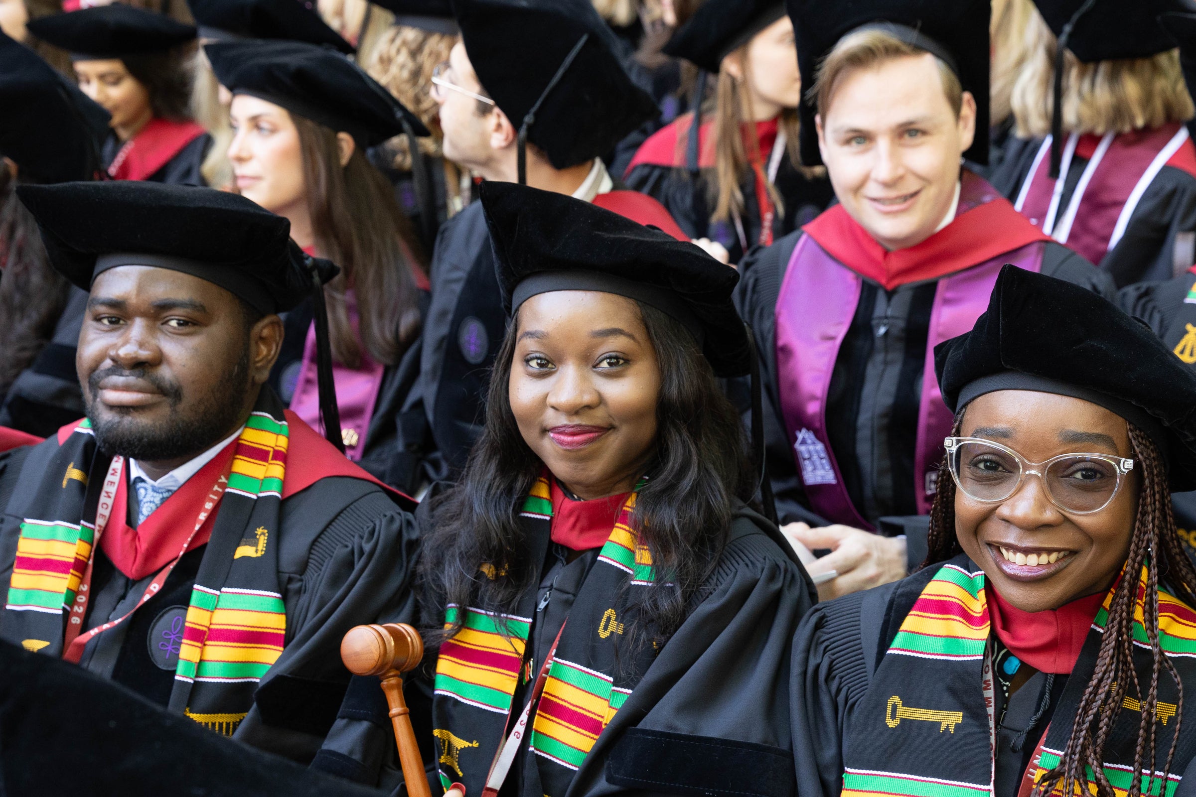 A group of graduates in regalia smile for the camera while seated for the ceremony.