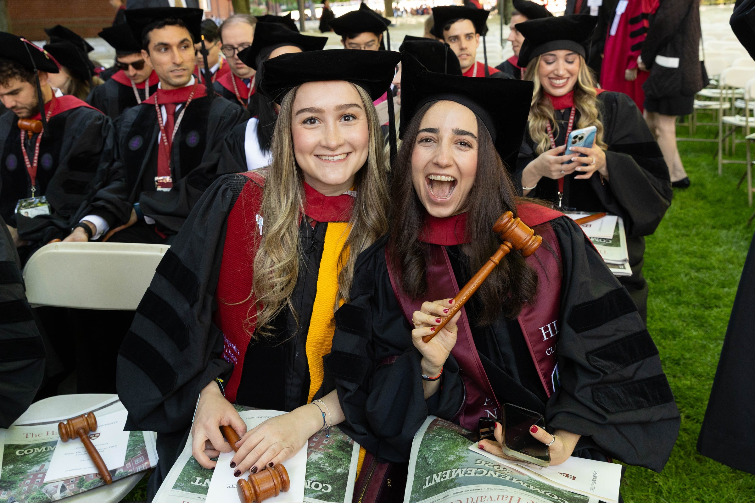 Two graduates in regalia smile for the camera while seated for commencement, one holding a wooden gavel.
