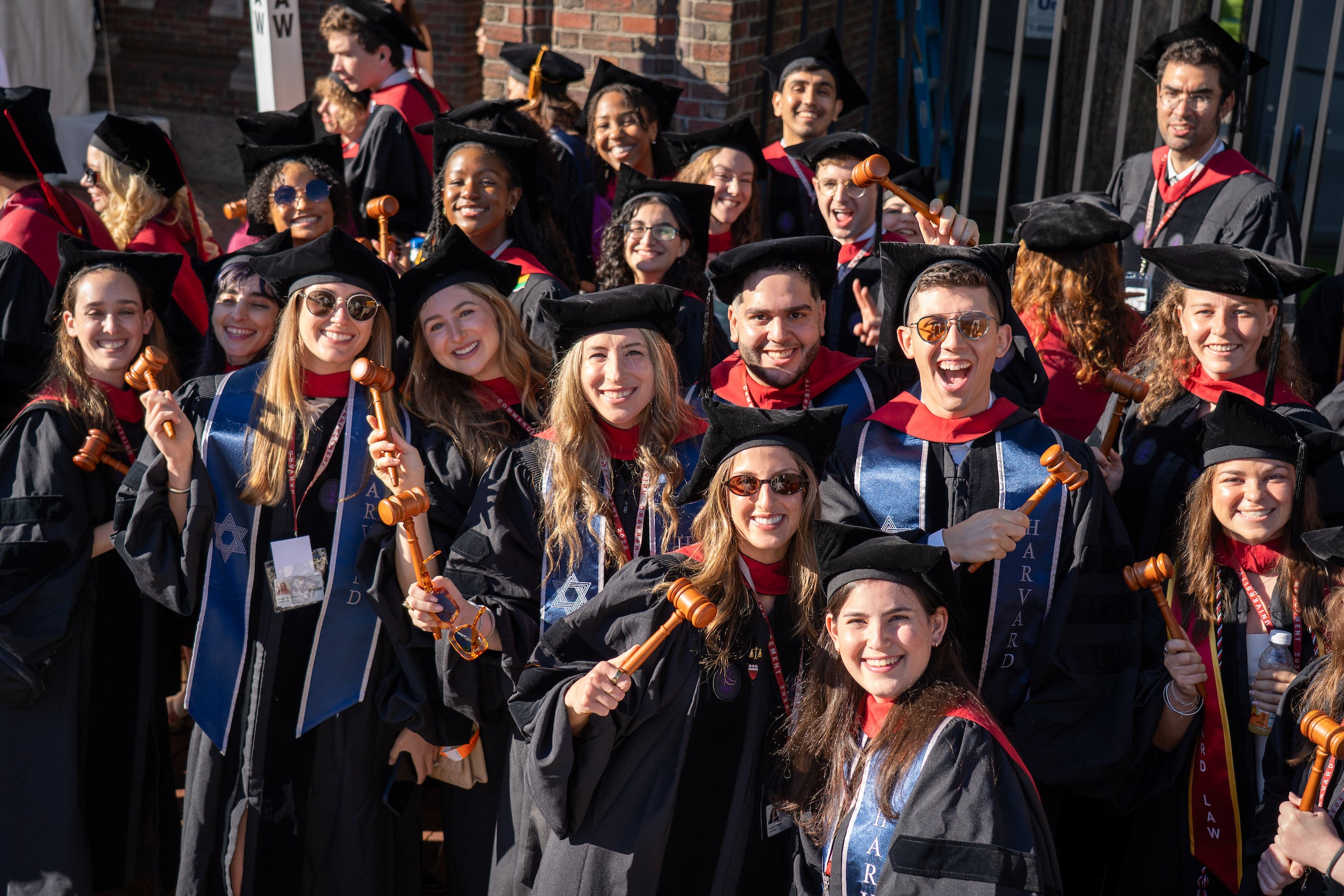 A group of graduates in black gowns and caps, some wearing blue sashes, pose together outdoors holding wooden gavels.