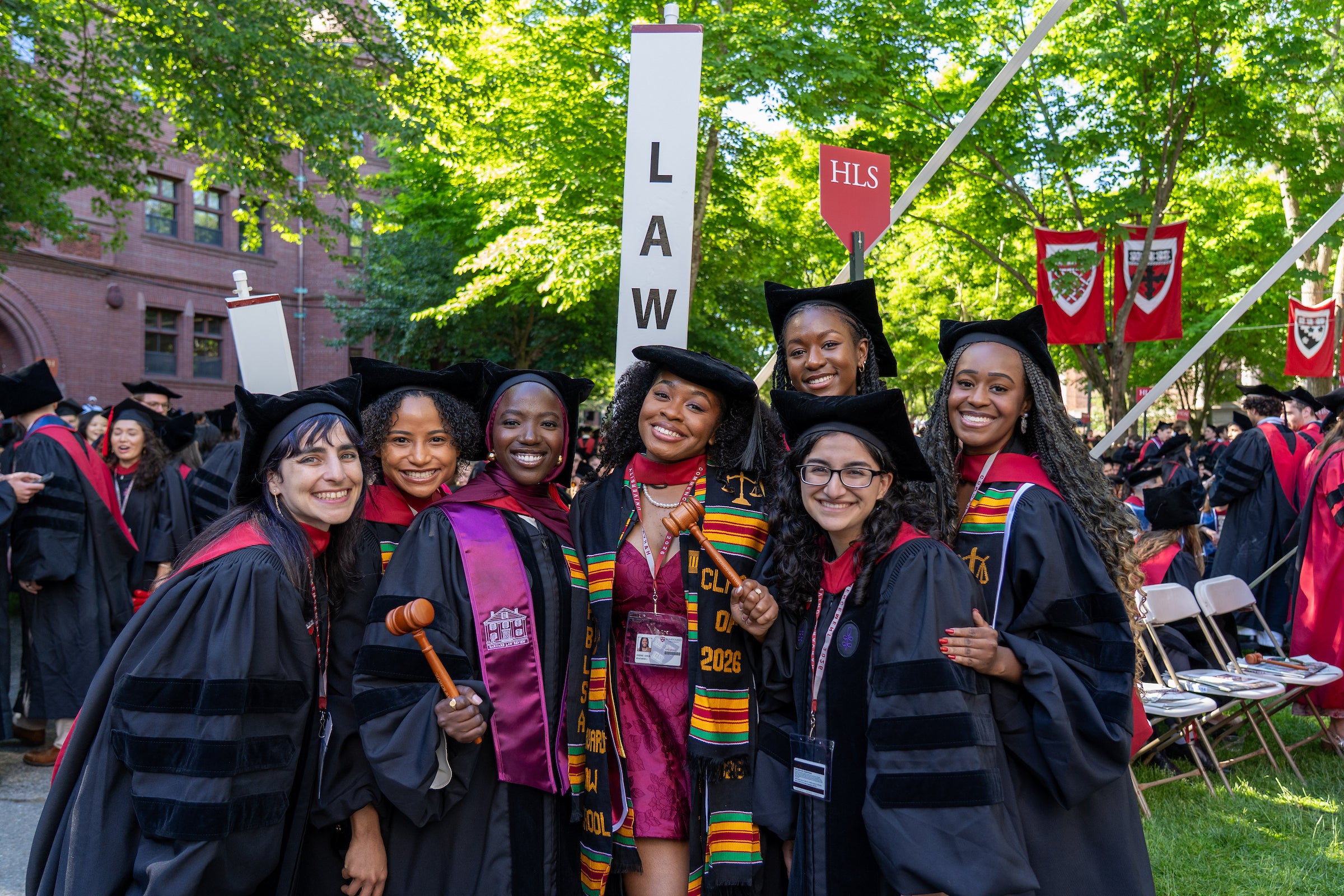 A group of graduates in black gowns and caps pose together outdoors at commencement, holding a gavel and wearing colorful stoles, with 