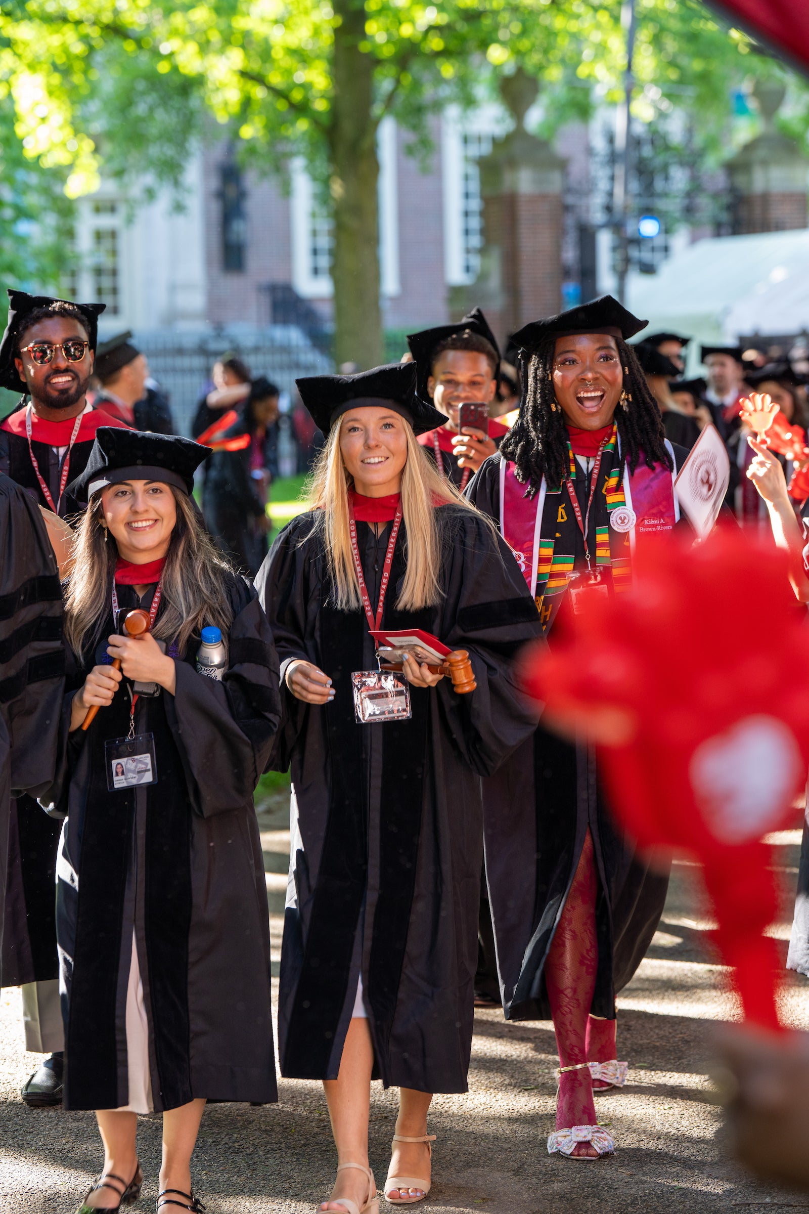 Graduates in black gowns and caps walk outdoors, smiling and holding diplomas, with trees and buildings in the background.