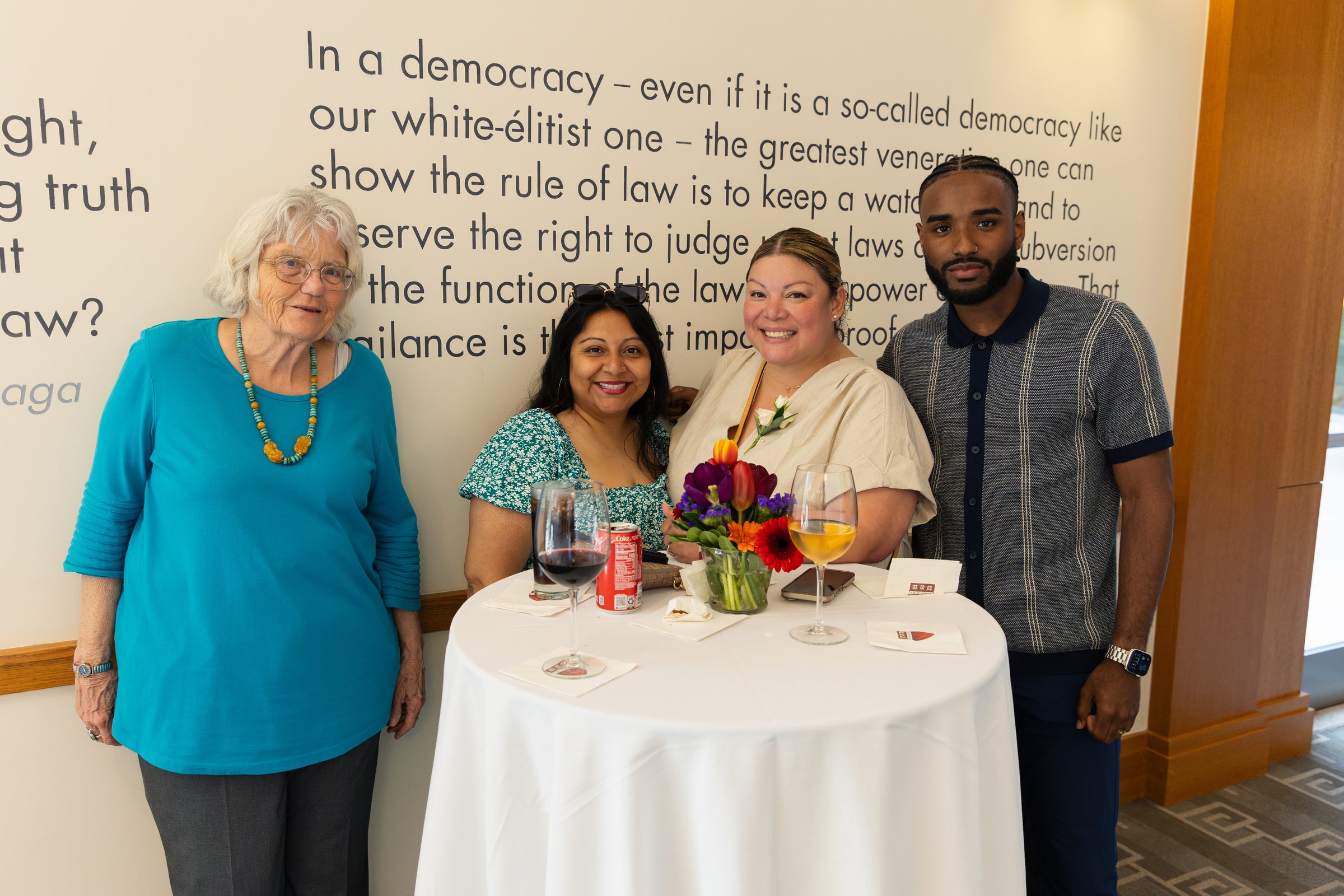 Four individuals pose together at a small table with drinks and flowers, standing in front of a wall with a large text quote.