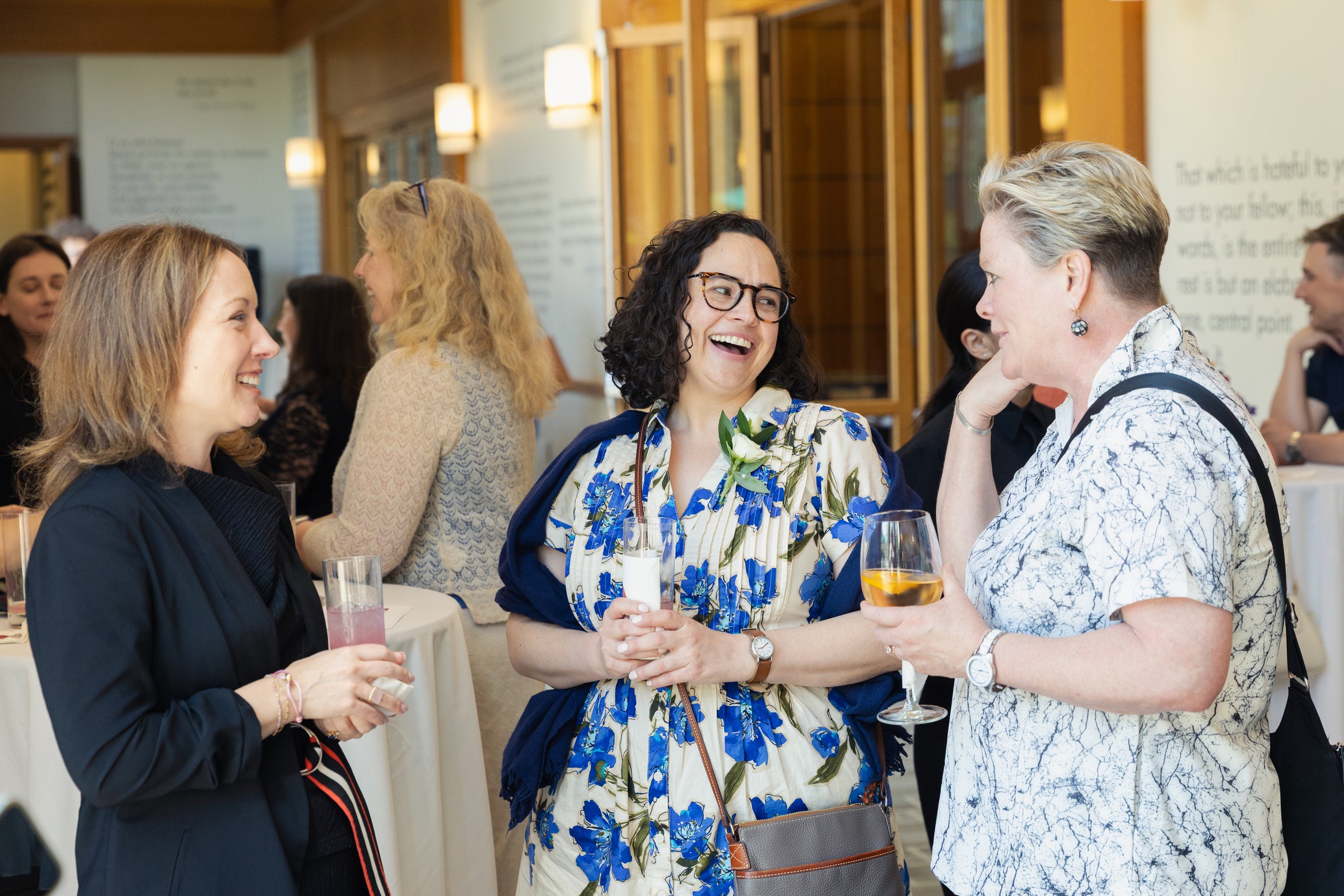 Three individuals laugh and chat while holding drinks at an indoor reception.