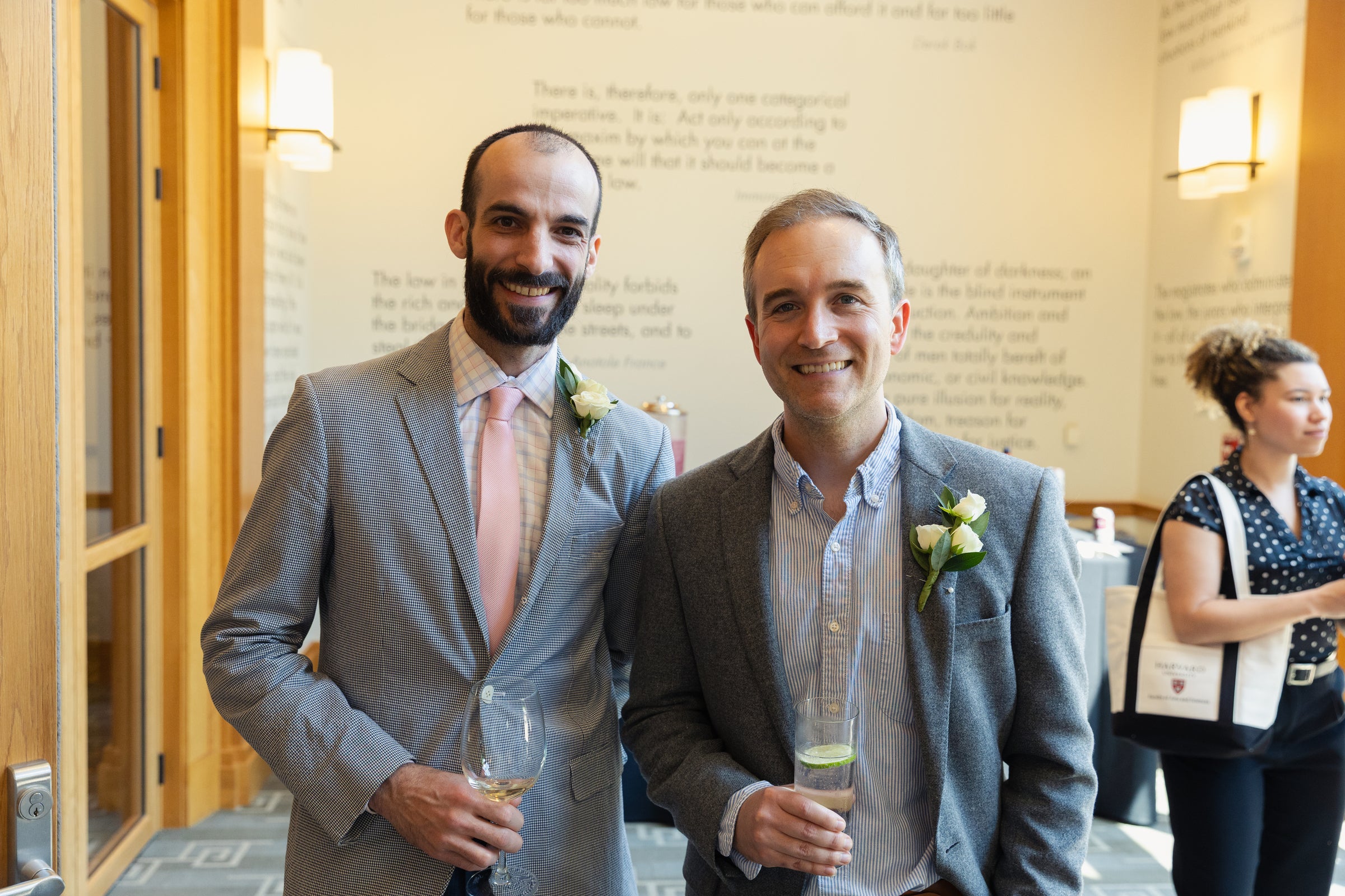 Two individuals in suits smile for the camera while holding drinks at an indoor event.